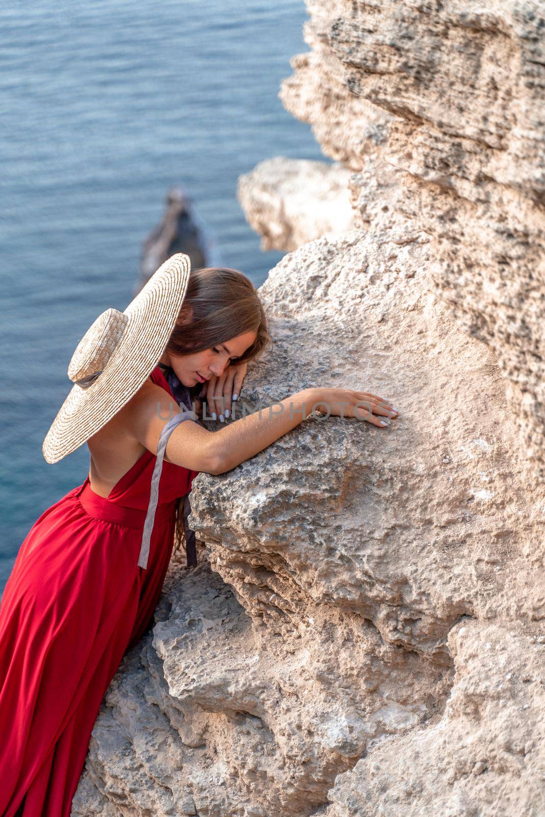 A girl with loose hair in a red dress and hat stands on the rocks above the sea. In the background, the sea and the rocks. The concept of travel