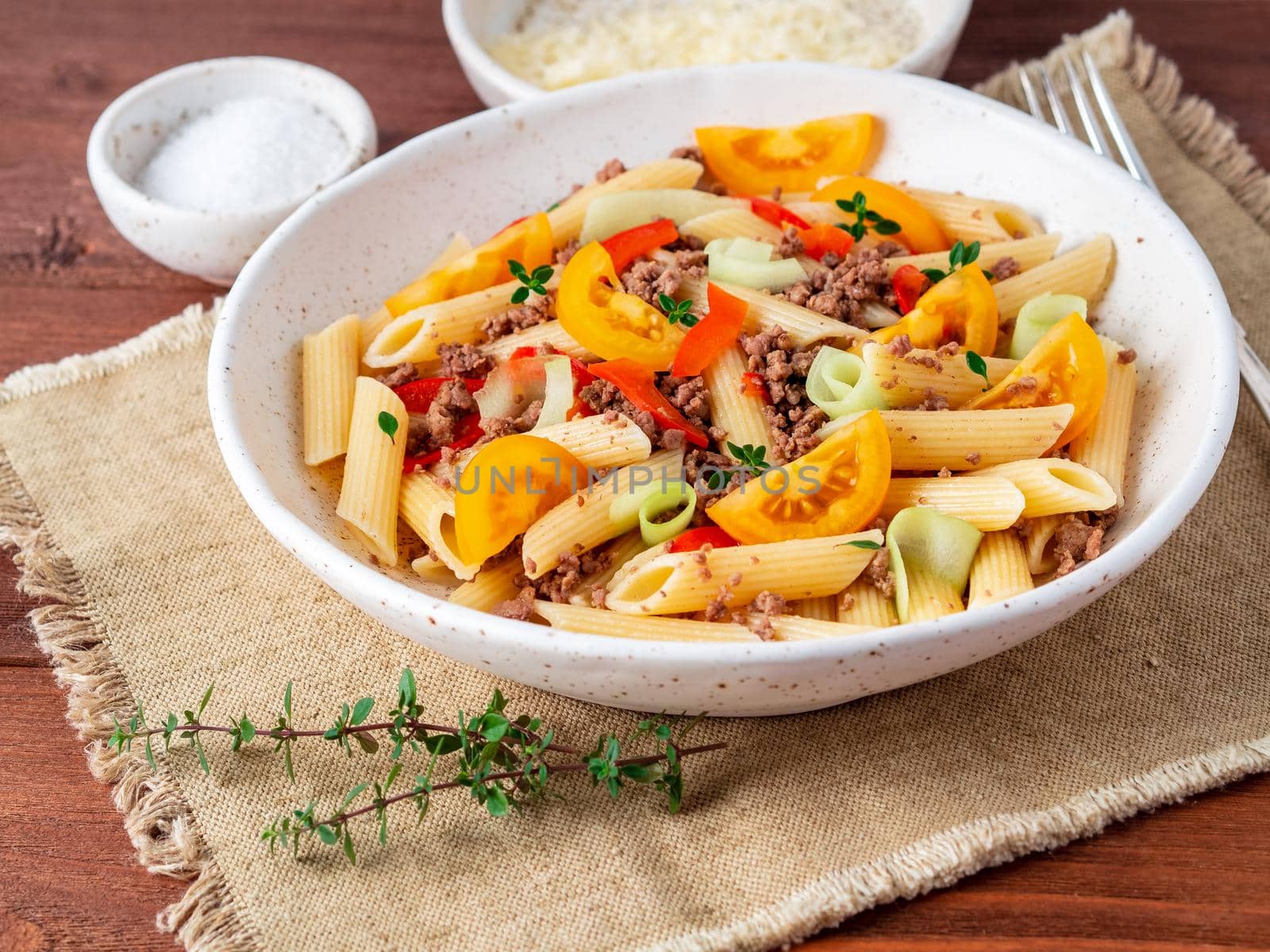 Penne pasta with yellow tomatoes, red and green vegetables, mincemeat on dark wooden background, side view.