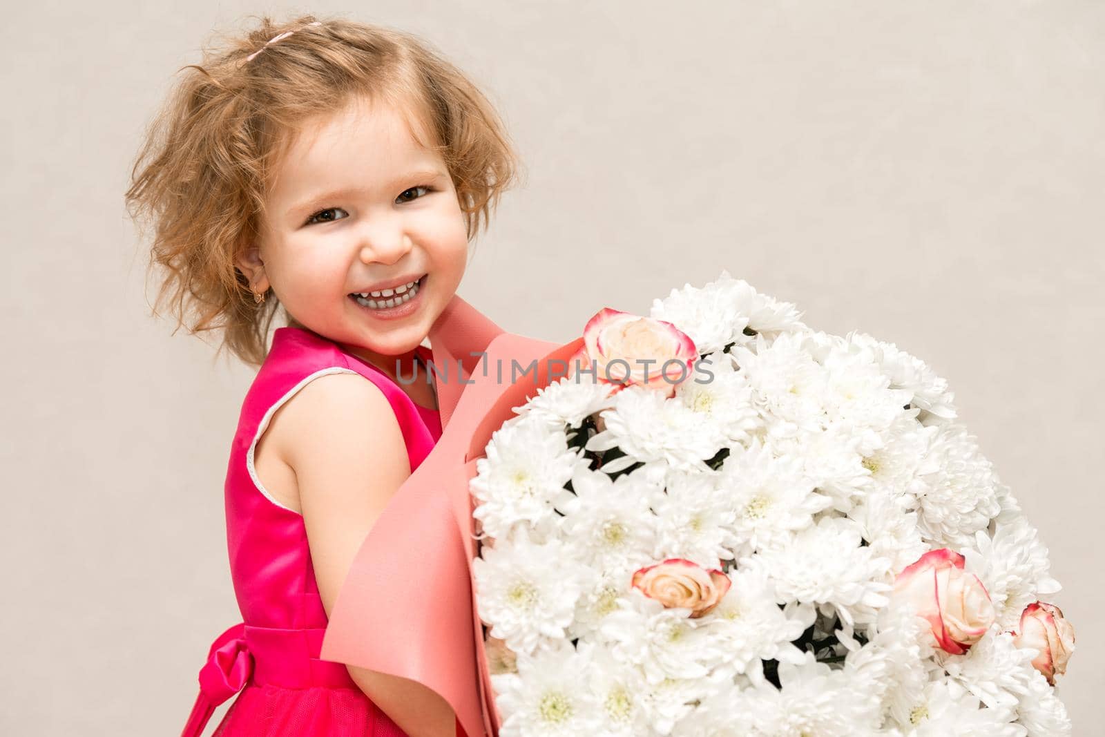 A little girl in a bright pink dress with curly hair is holding a huge bouquet of chrysanthemums and roses for her mother and smiling broadly and fervently. by olex