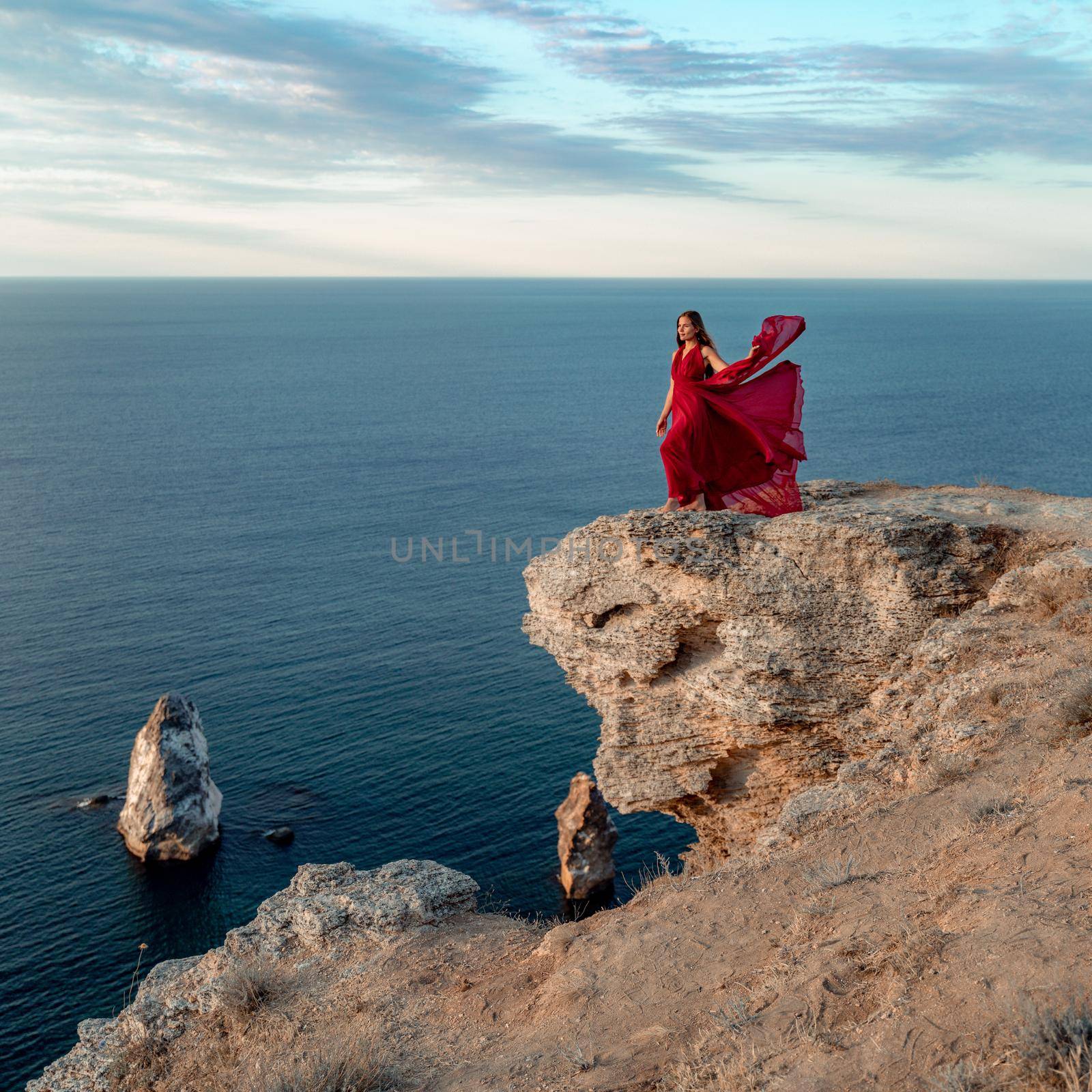 A girl with loose hair in a red dress stands on a rock rock above the sea. In the background, the sea and the rocks. The concept of travel by Matiunina