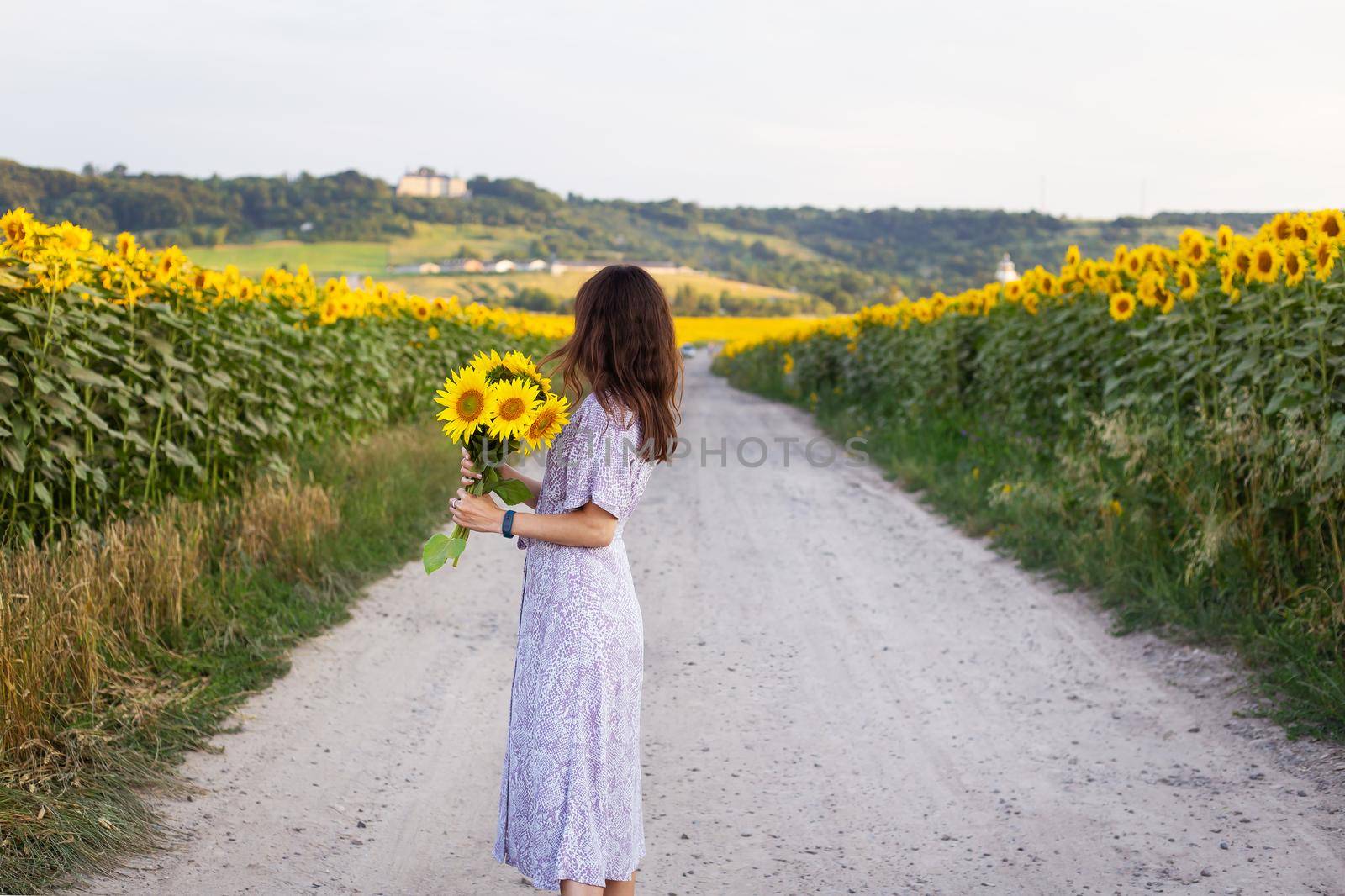 A beautiful young girl in a dress holds a bouquet of sunflowers in her hands stands on the road among a large field with sunflowers