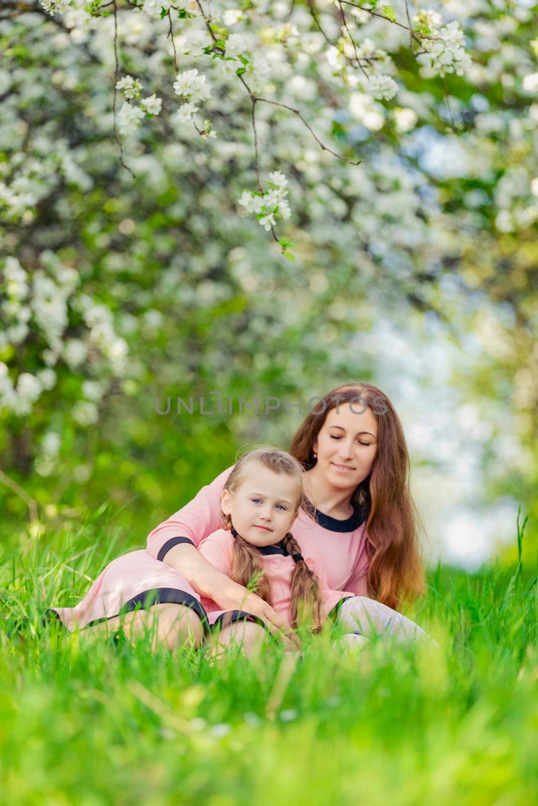 mother and daughter sit in the green grass against the backdrop of blooming apple trees