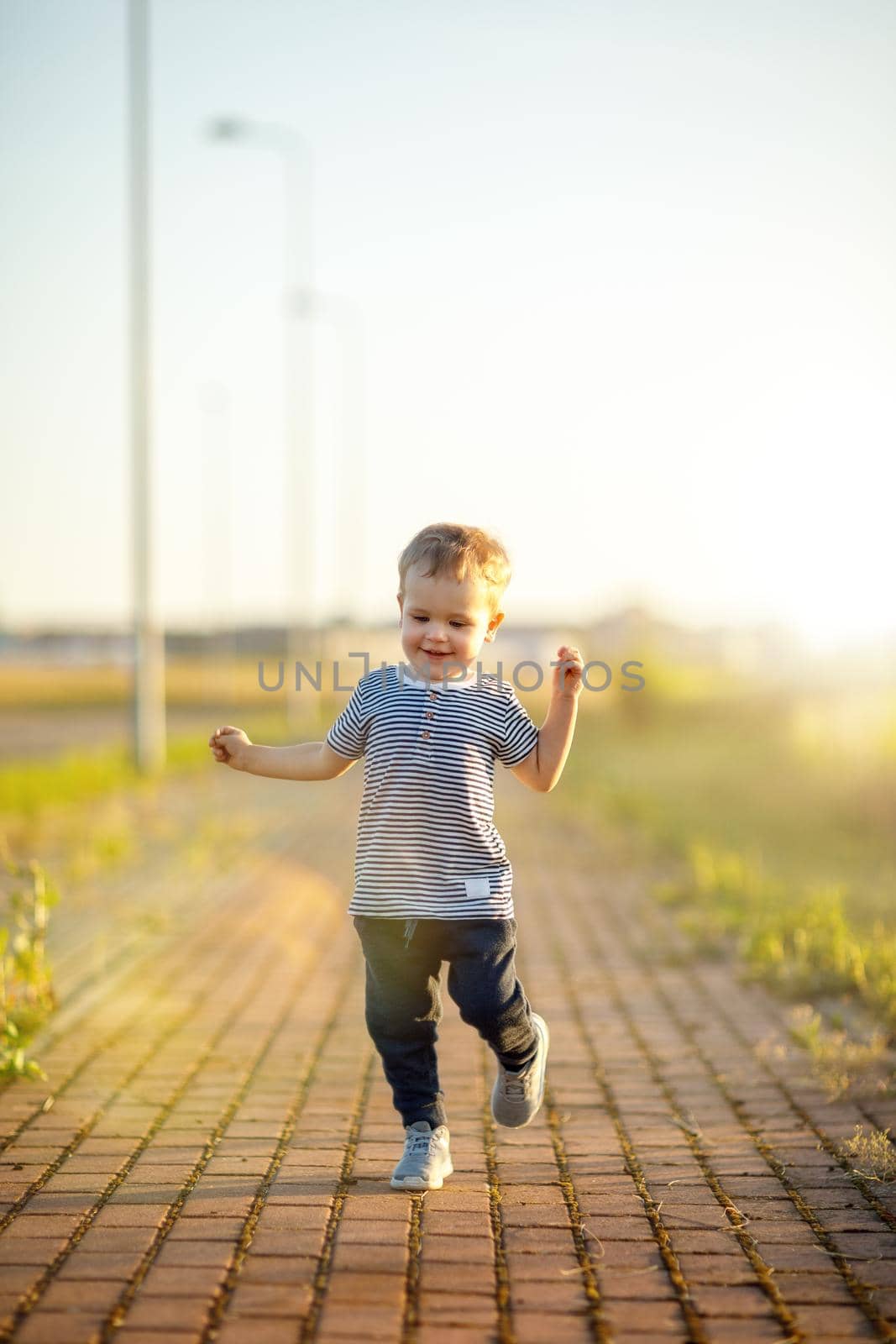 Cheerful little boy with striped shirt runs down the sidewalk. Sunny summer evening, backlighting.