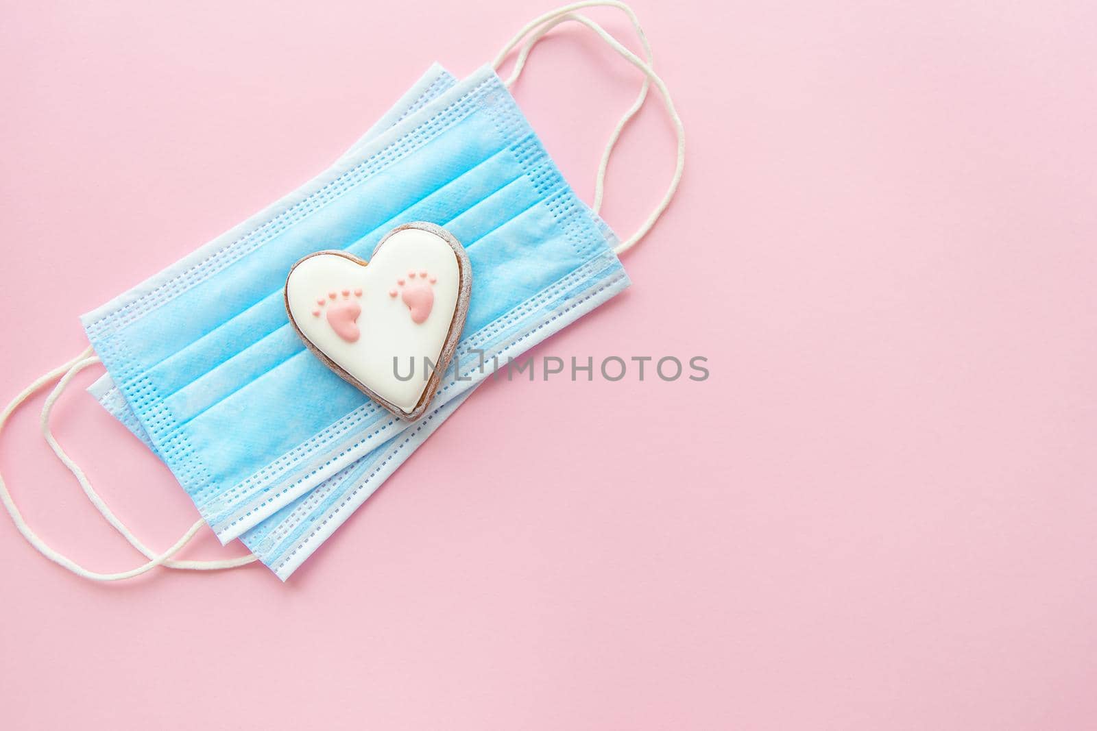 Disposable medical mask and heart with baby footprint on pink background. Childbirth during the coronavirus pandemic