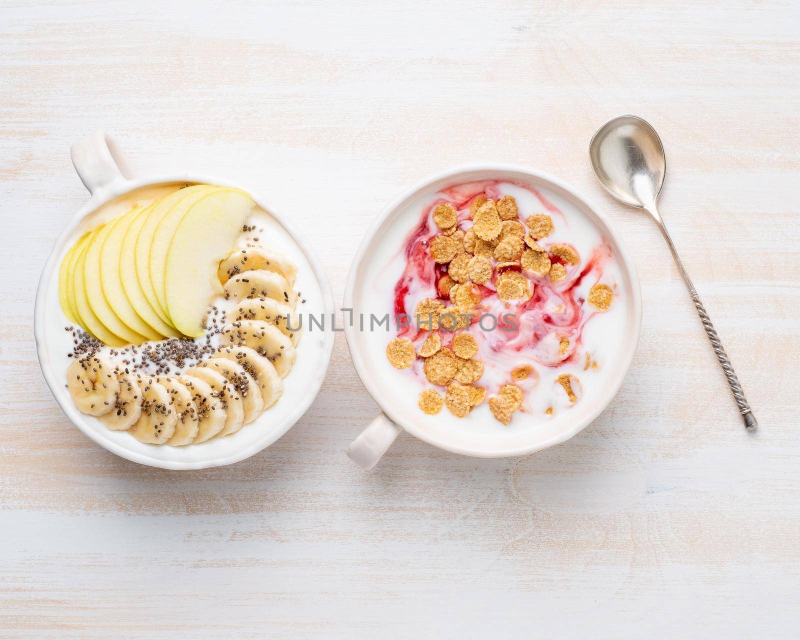 Two greek yogurt with jam, apple, muesli, chia seeds and banana in white bowl on white wooden table, top view.