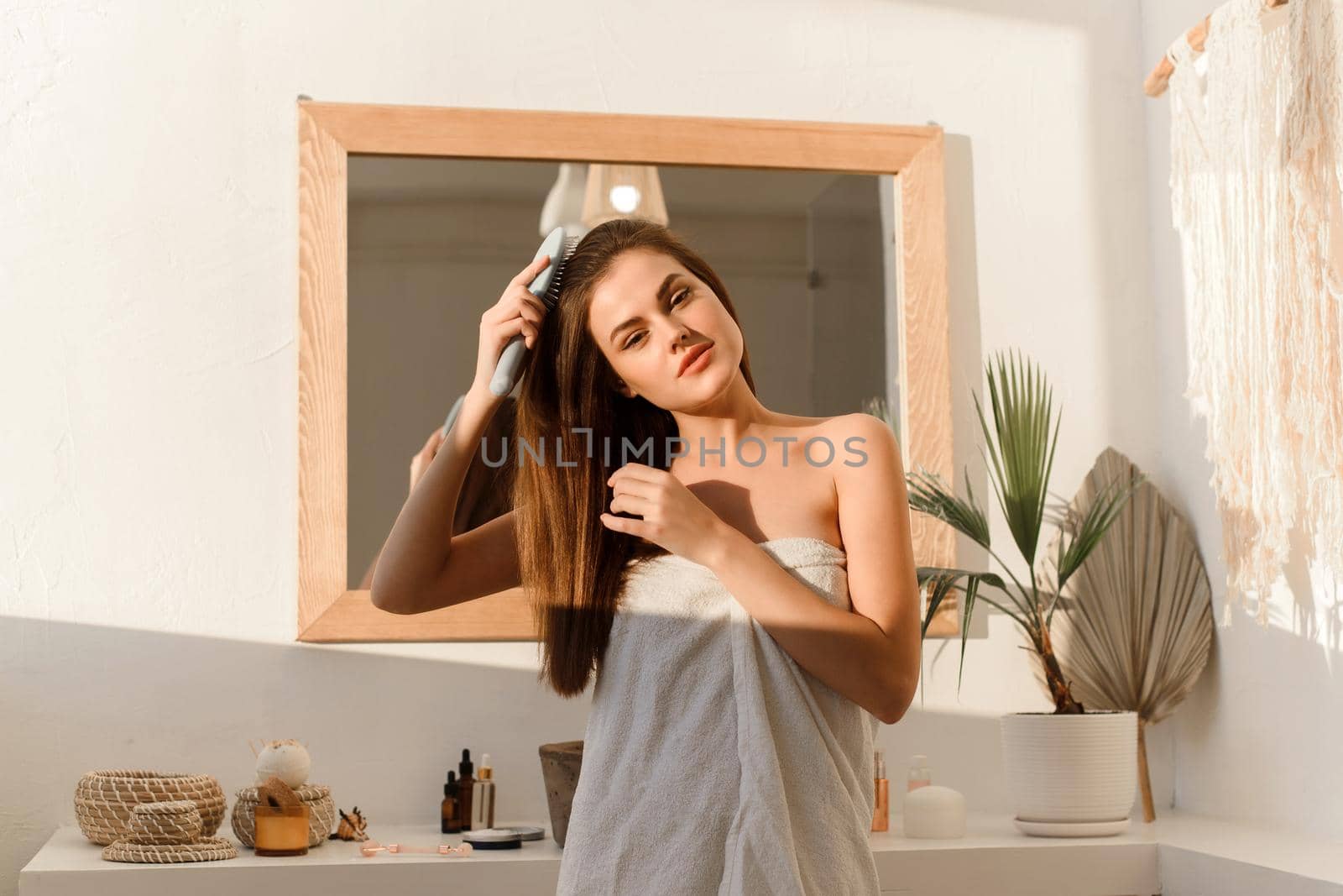 Smiling pretty brunette in a towel combs her long shiny silky hair with a comb in the morning after cosmetic procedures, on the background of the bathroom