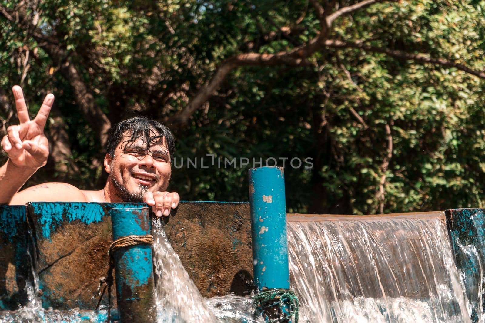 Latino man smiling and looking at the camera making the peace gesture with his hand and fingers submerged in a river dam in Masachapa, Nicaragua. Photo with copy space.