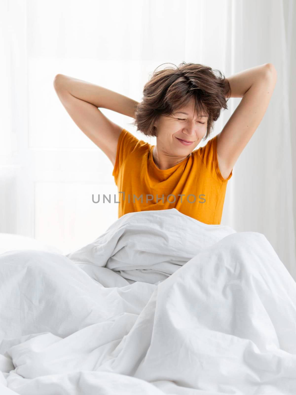 Sleepy woman in yellow pajama is stretching in bed, covered with white blanket. Waking up early in morning. Woman gets enough sleep.