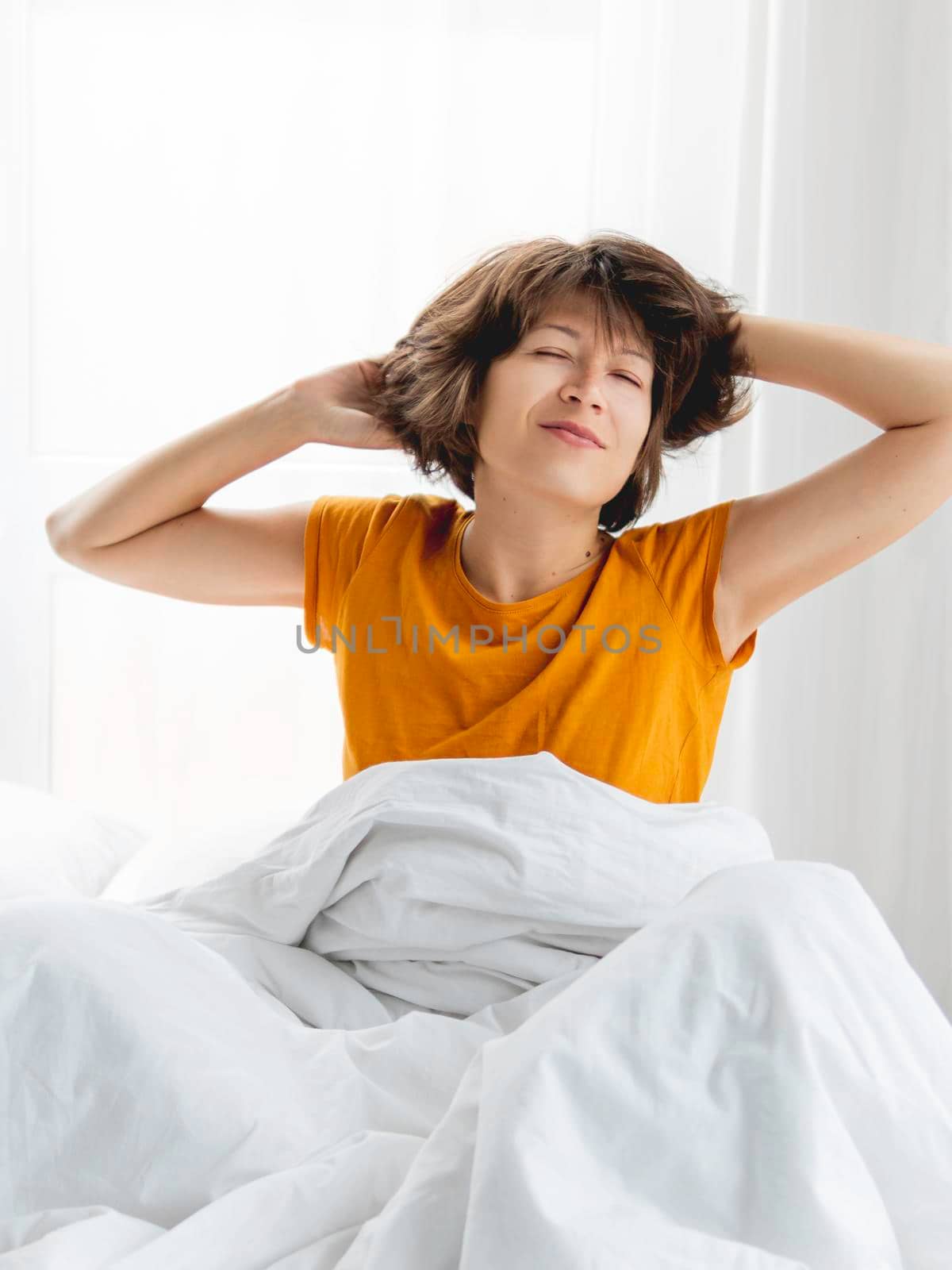 Sleepy woman in yellow pajama is stretching in bed, covered with white blanket. Waking up early in morning. Woman gets enough sleep. by aksenovko