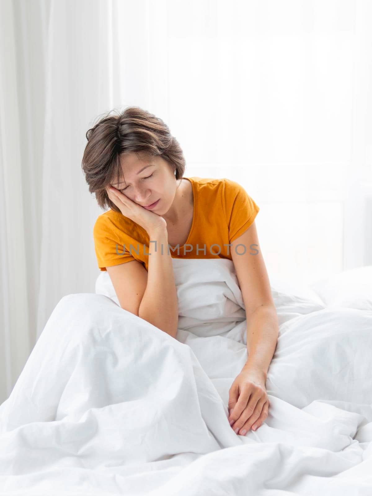 Sleepy woman is sitting in bed, completely covered with white blanket. It's hard to wake up early in morning. Woman does not get enough sleep.