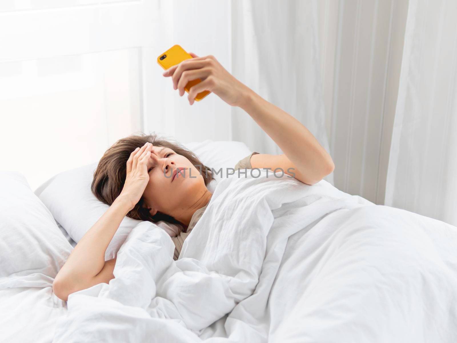 Sleepy woman looks on smartphone screen. Checking e-mail box right after waking up. Using wireless technology in bed. by aksenovko