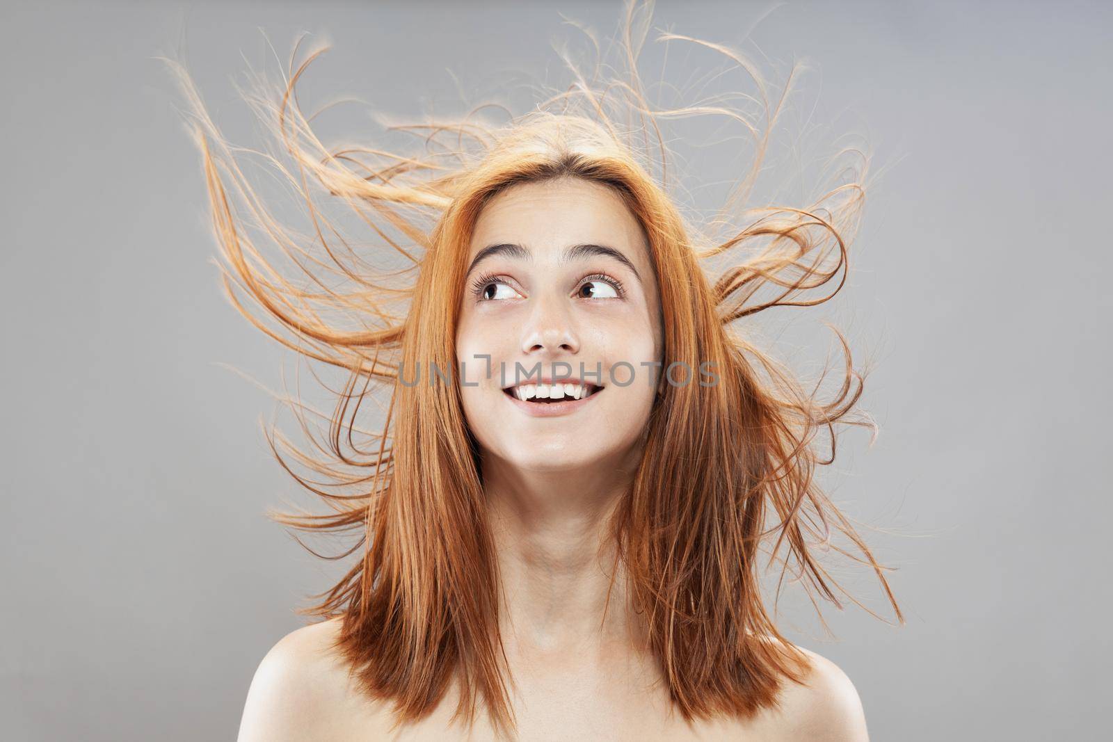 Beautiful dark burnt orange windy hair girl smiling. Studio portrait with happy face expression against gray background... by kokimk