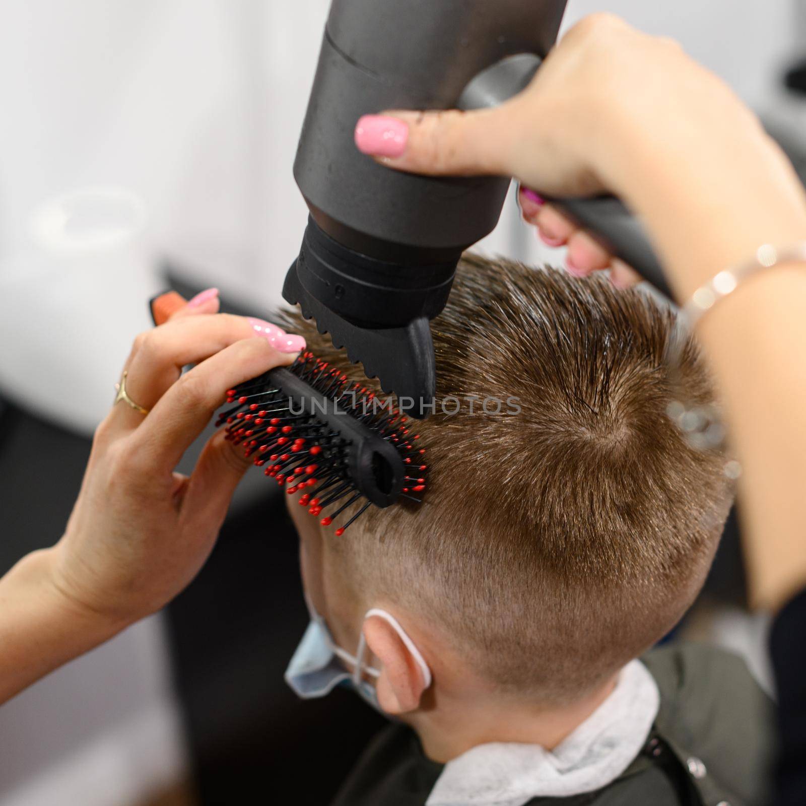 The hairdresser dries and styles the hair with the help of a hair dryer and a comb for a schoolboy boy, visits to the hairdresser during a coronavirus pandemic, black style hairdresser.