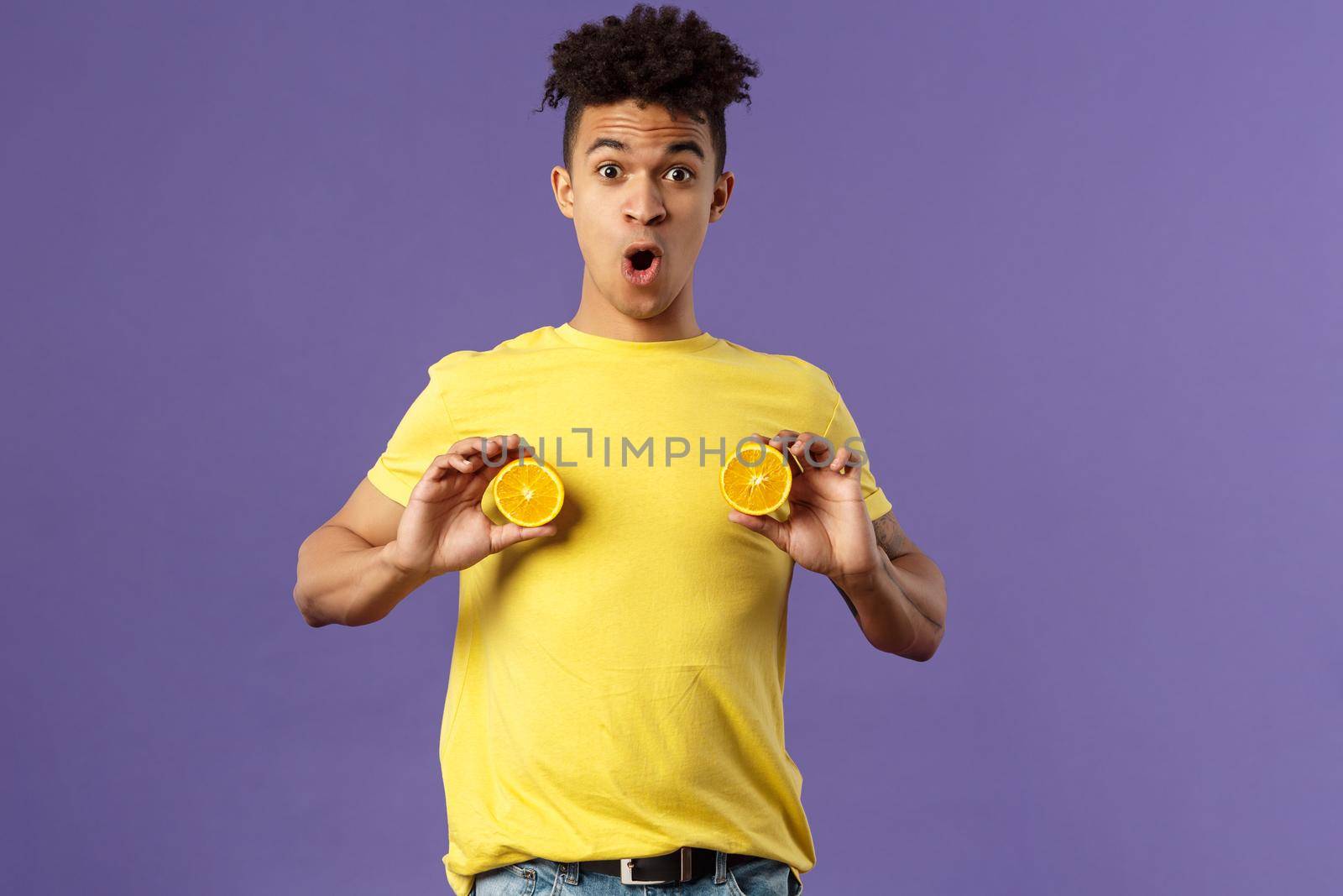 Holidays, vitamins and vacation concept. Portrait of funny and cute young 25s man fool around, showing breast with pieces of oranges over chest, look ashamed or shocked, purple background.