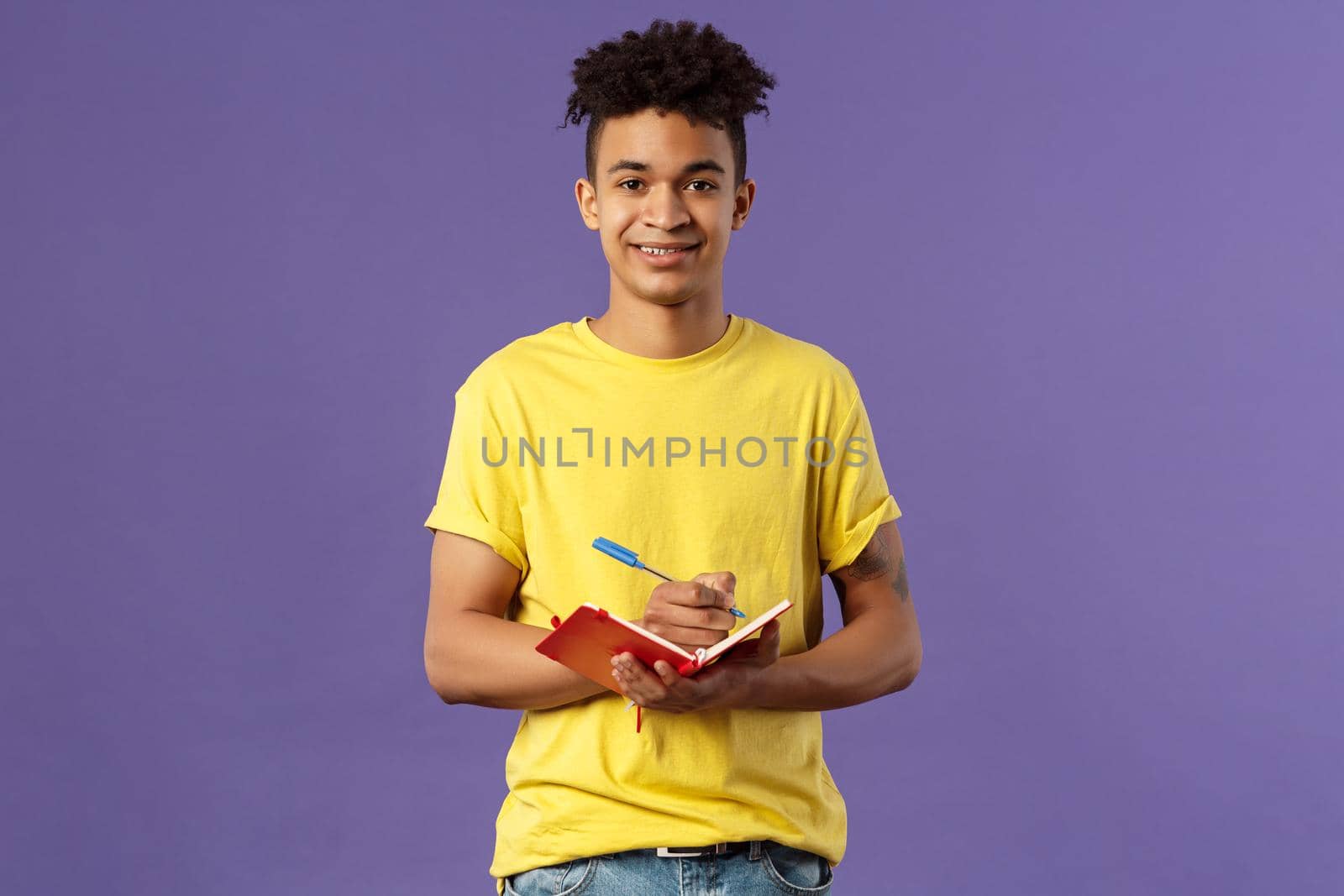 Portrait of young hispanic male student studying online courses, writing down lecture, making personal schedule or taking notes in planner, look camera enthusiastic and interested, purple background.