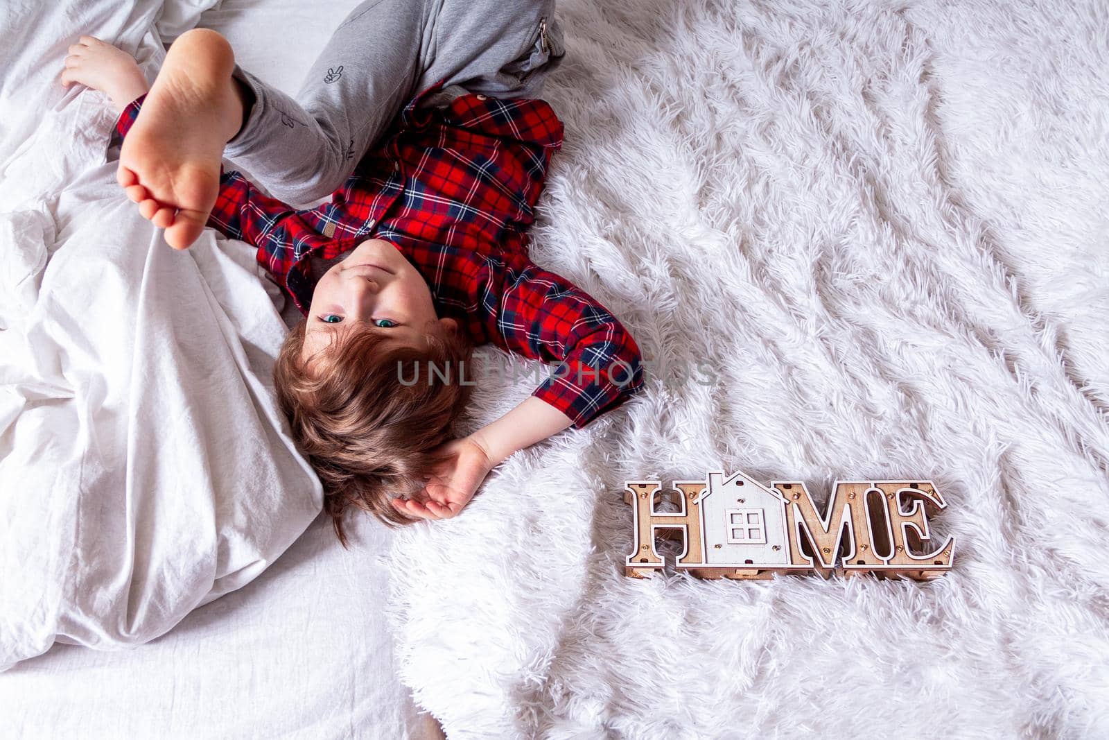 Happy little curly kid boy with smiley face lying on the white bed at home and legs up. Stay at home quarantine coronavirus COVID-19 pandemic prevention. With space for text and wooden house