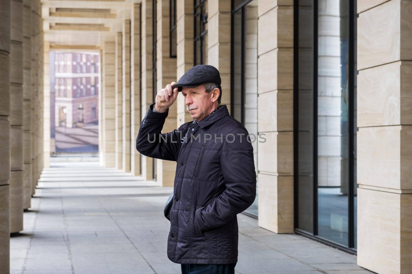 Adult Caucasian male stands near modern building with columns. Pensioner in seasonal clothes while walking around the city. Selective focus.