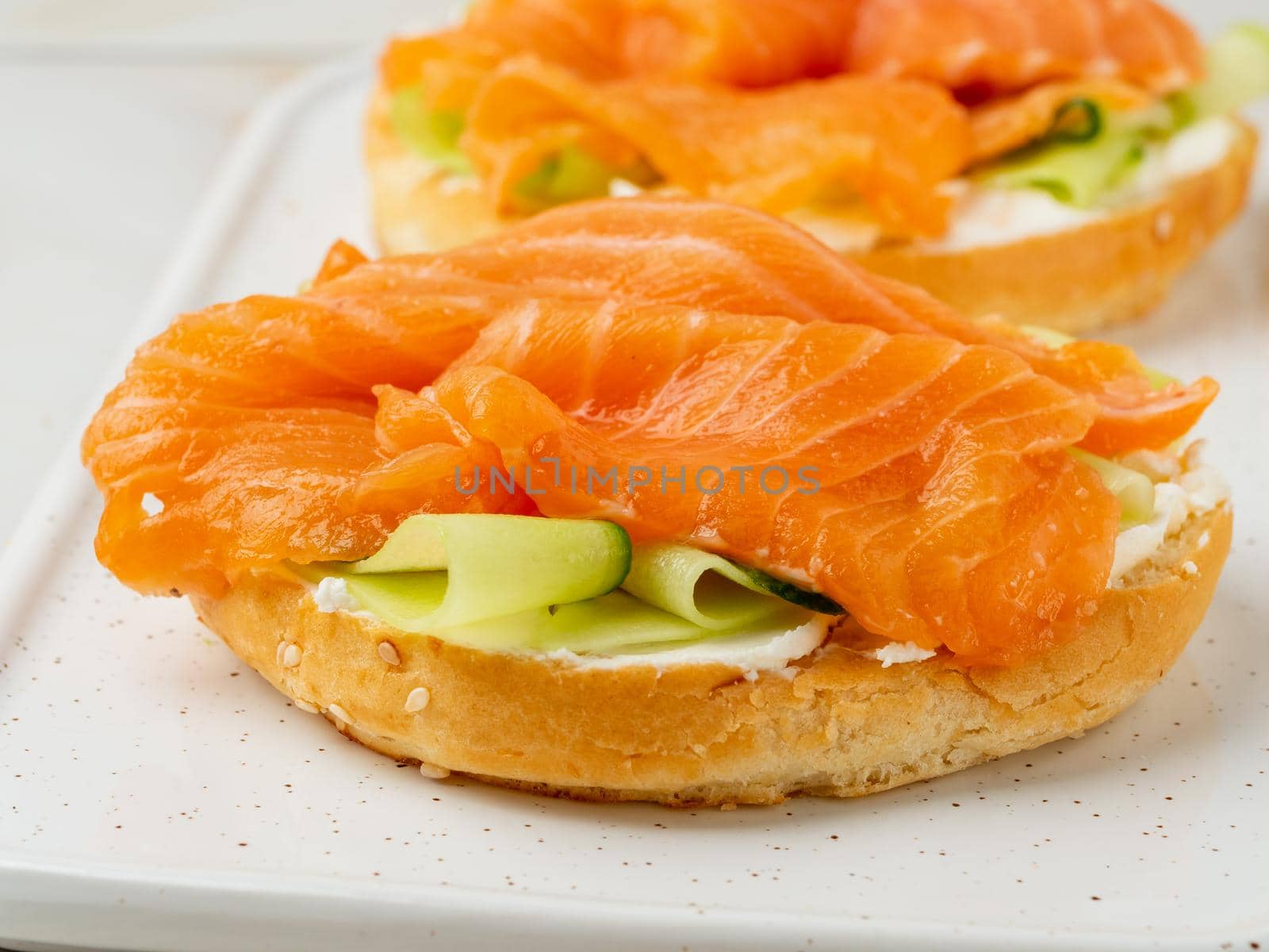 Two open sandwiches with salmon, cream cheese, cucumber slices on white marble table, close up
