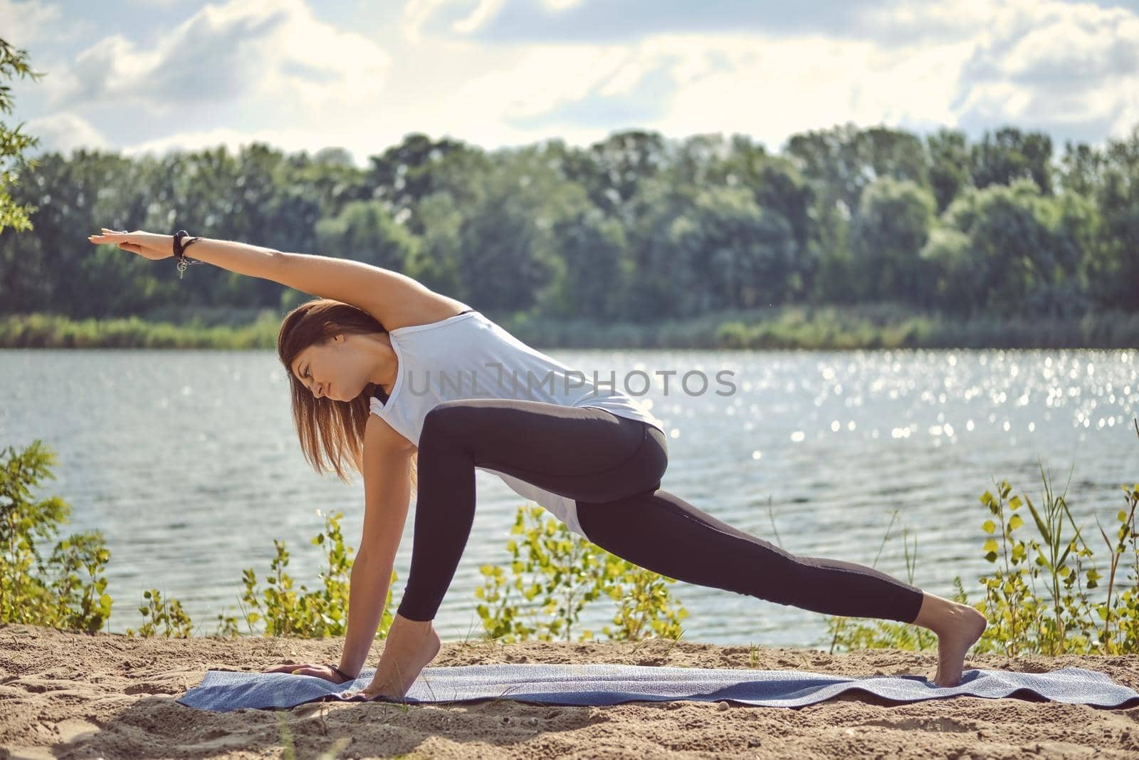 Graceful dark-haired maiden in white undershirt and black leggings is practicing yoga performing yoga-asanas and doing stretching on a mat laying on a sand near a riverside. Concept of healthy life and natural balance between body and mental development.