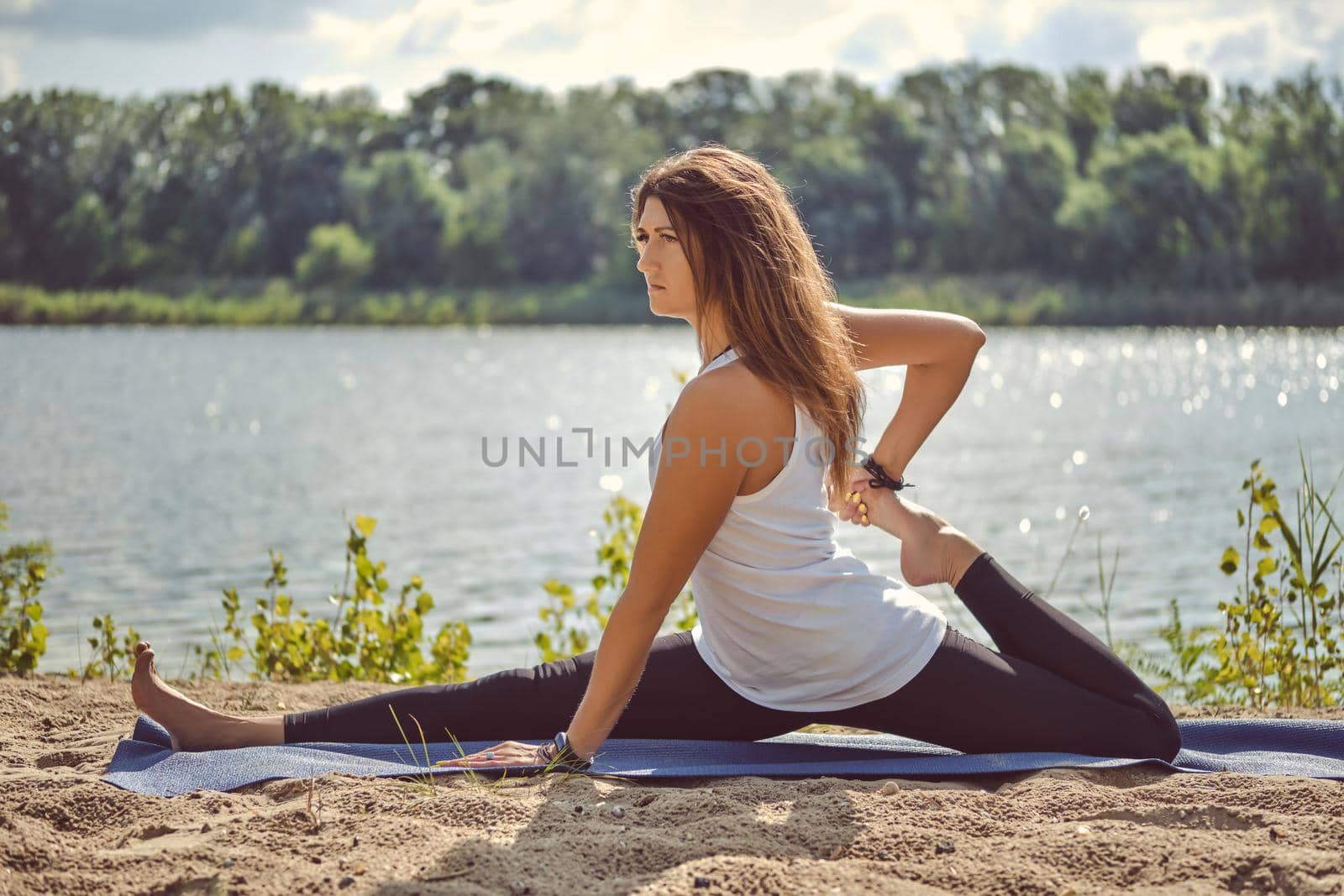 Pretty dark-haired girl in white undershirt and black leggings is practicing yoga performing yoga-asanas and doing stretching on a mat laying on a sand near a riverside. Concept of healthy life and natural balance between body and mental development.