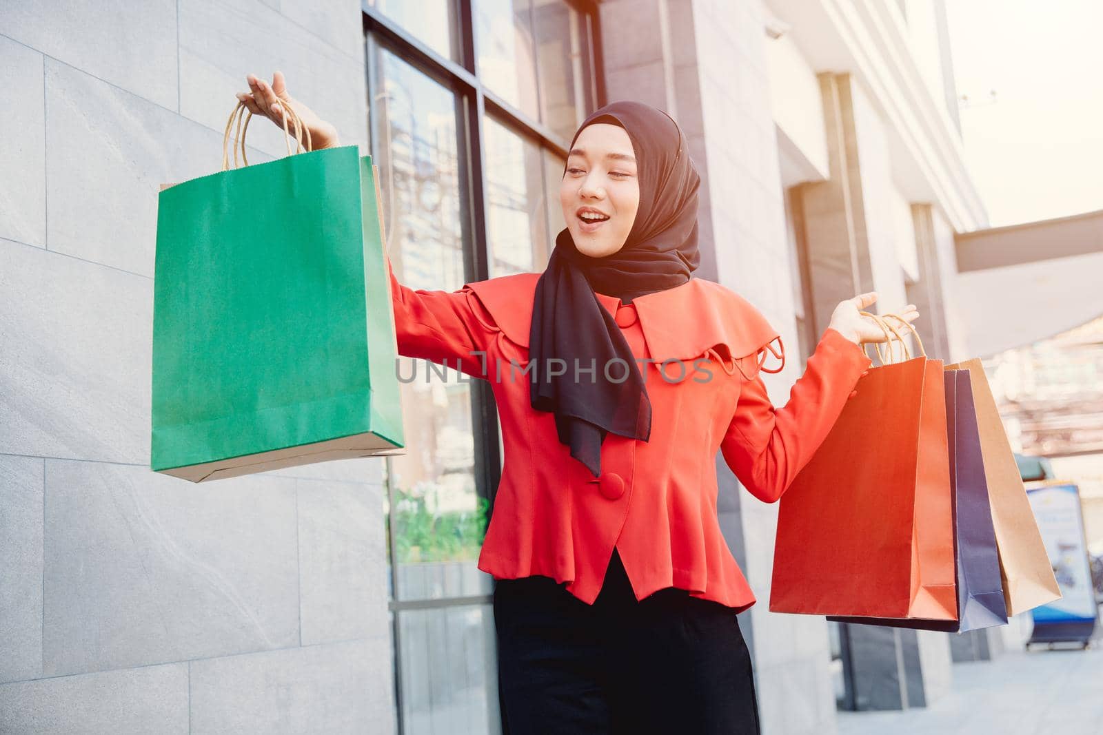 Arab Muslim young woman in veil hijab clothes holding shopping bags and walking on the city street. Shopping time. Modern skyscrapers in the background. by qualitystocks