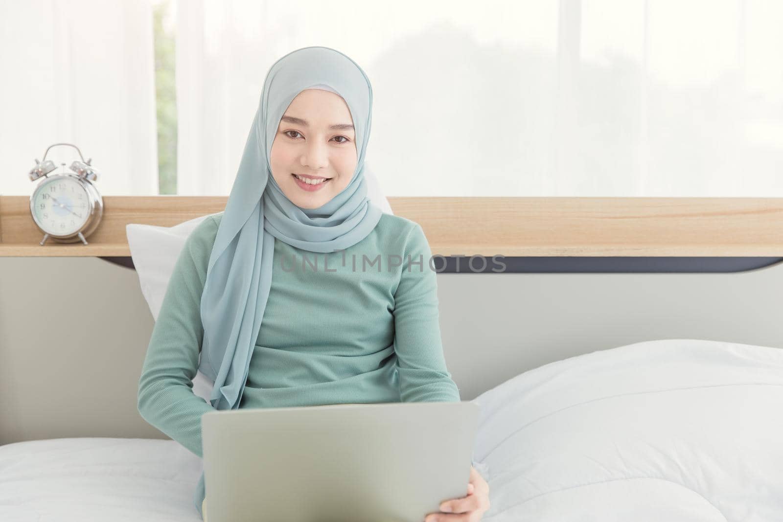 Asian Muslim Islamic woman with hijab happy enjoy online remote work at home with her laptop on the bed. by qualitystocks