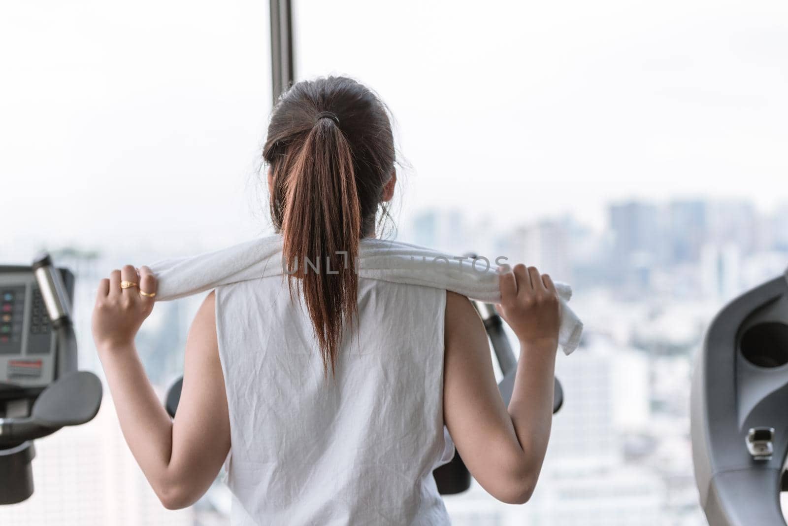 Asian woman morning exercise in sport club with towel on her shoulders looking city at the windows view from behind.
