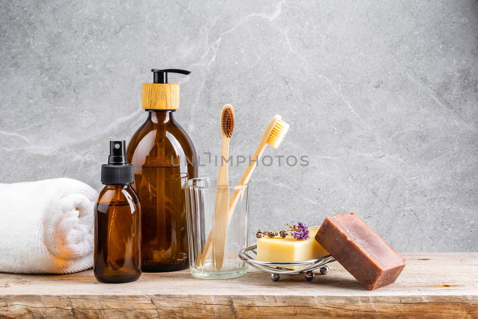 Bathroom table with white towel, dark glass bottles with liquid skin care products, bamboo tooth brushes and natural handmade solid soap bars over grey marble background