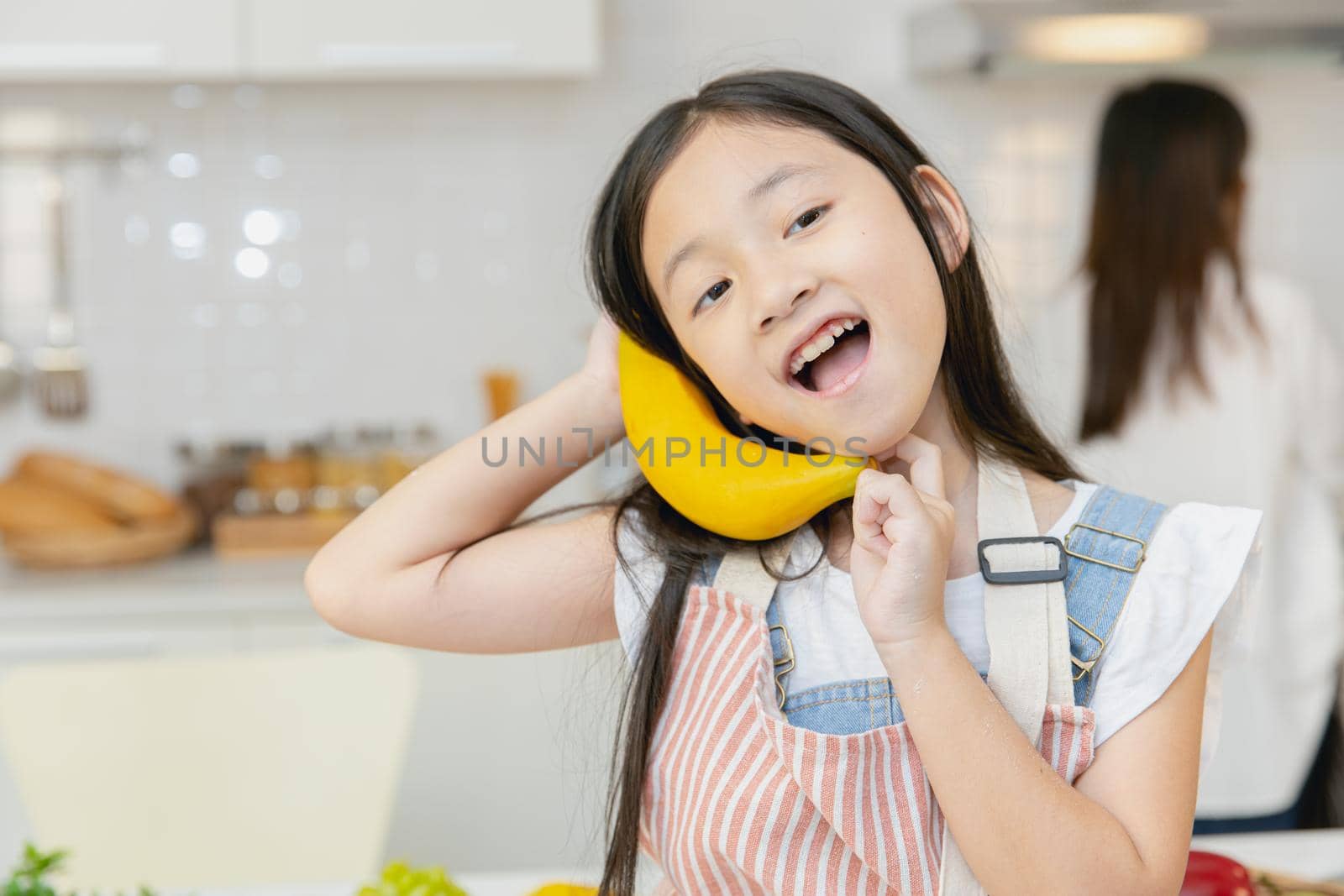 Cute girl child enjoy funny candid moment playing with banana at home kitchen.