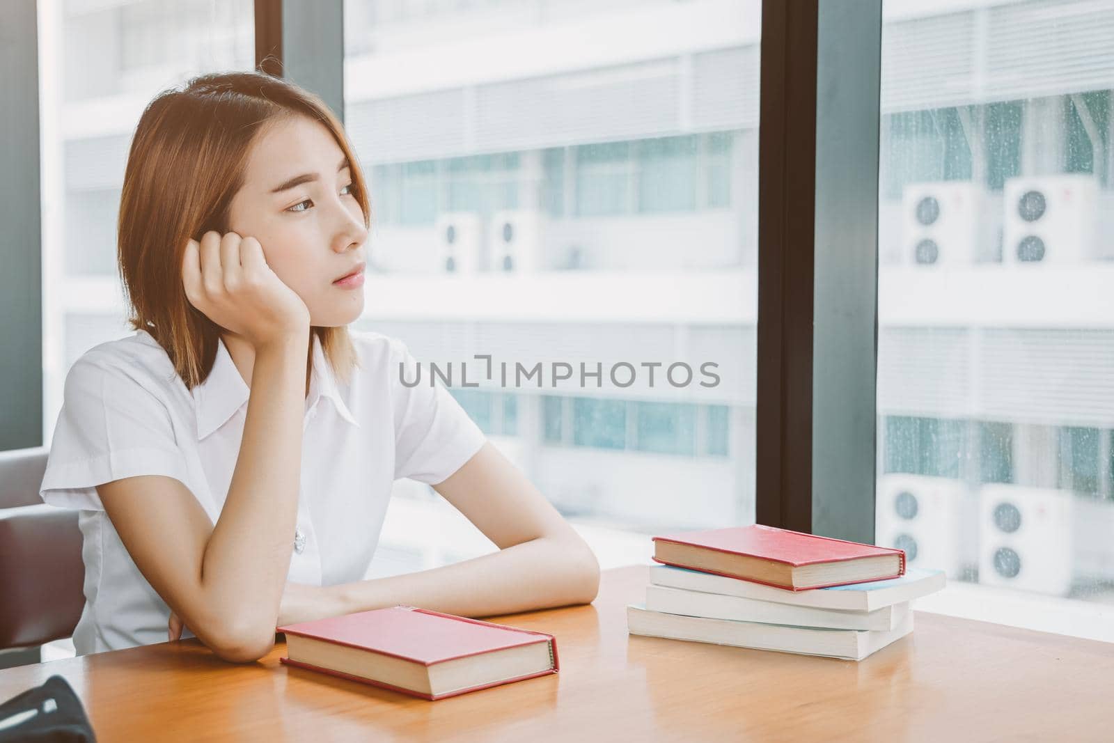Asian university girl teen sitting alone thinking worry, day dreaming or boring looking out window lonely expression in library.