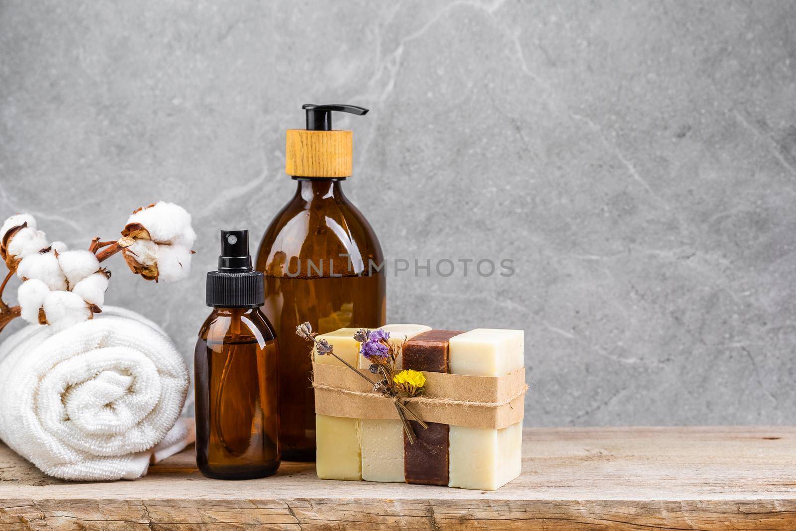 Bathroom table with white towel and cotton flower branch, dark glass bottles with liquid skin care products and natural handmade solid soap bars in stack over grey marble background