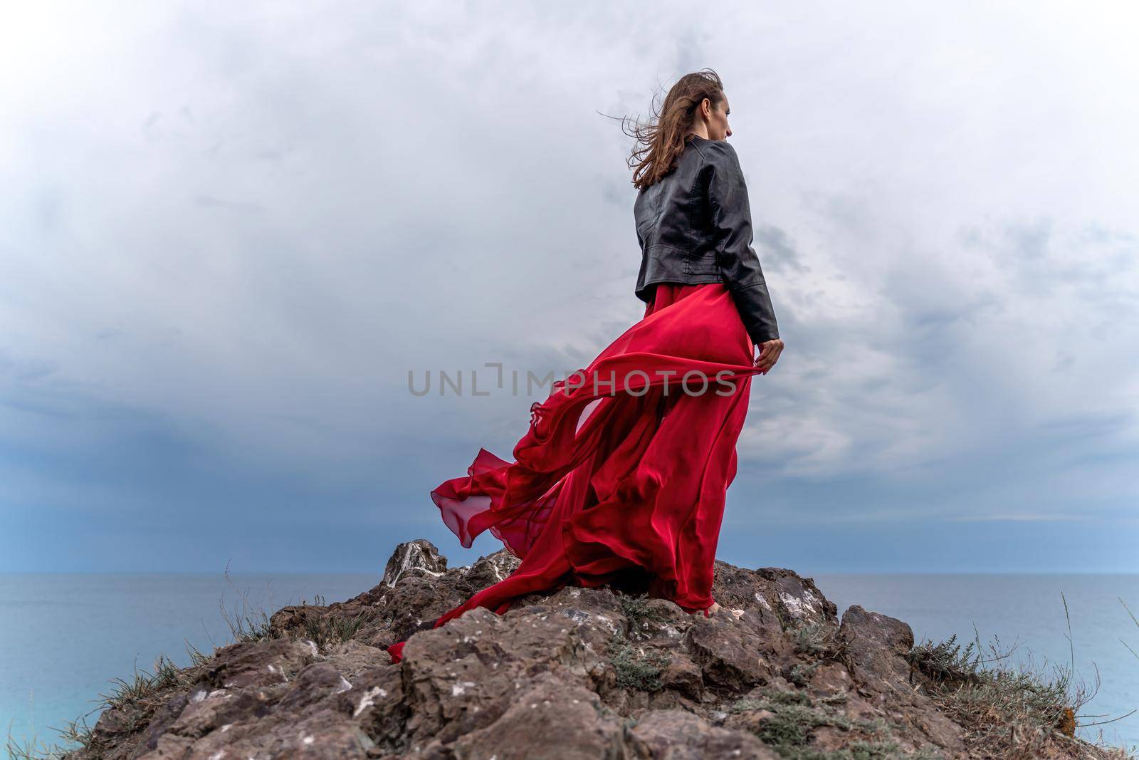 A woman in a red dress and leather jacket stands above a stormy sky, her dress fluttering, the fabric flying in the wind