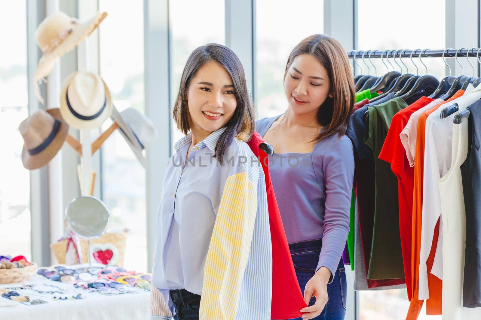 happy women shopping with friend in fashion shop for select new colorful clothes and measure size at shoulder.