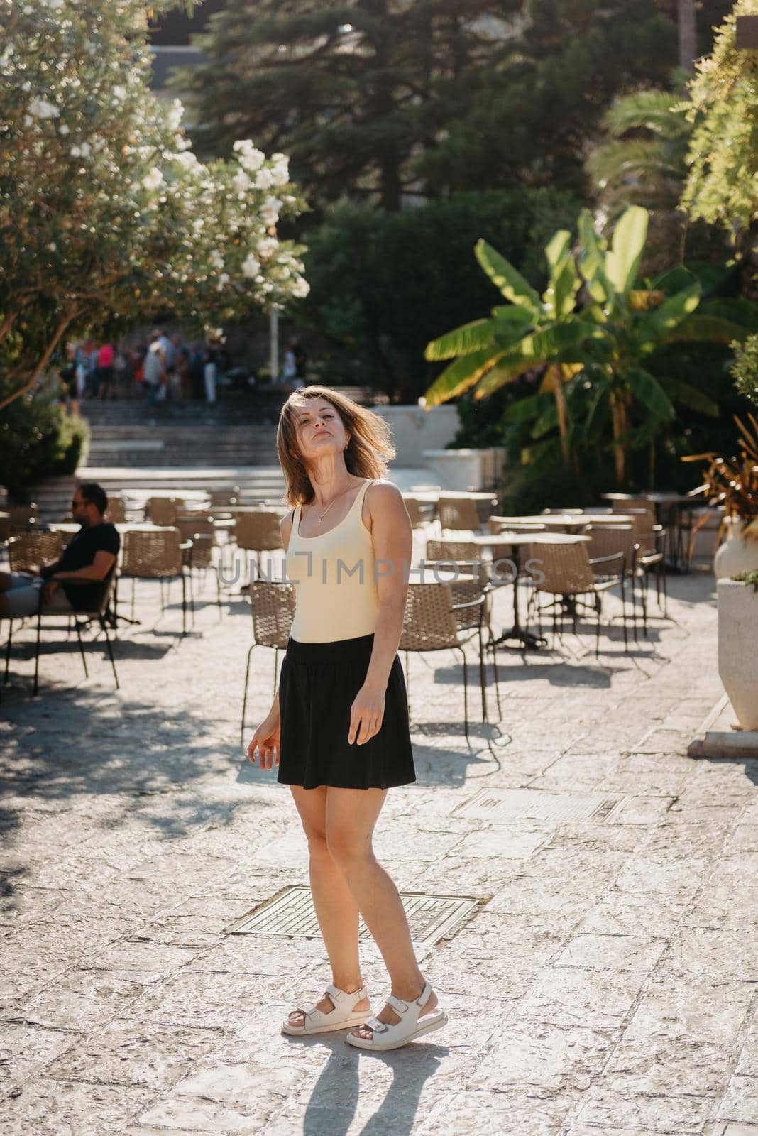 Girl Tourist Walking Through Ancient Narrow Street On A Beautiful Summer Day In MEDITERRANEAN MEDIEVAL CITY, OLD TOWN BUDVA, MONTENEGRO. Young Beautiful Cheerful Woman Walking On Old Street At Tropical Town. Pretty Girl Looking At You And Smiling by Andrii_Ko