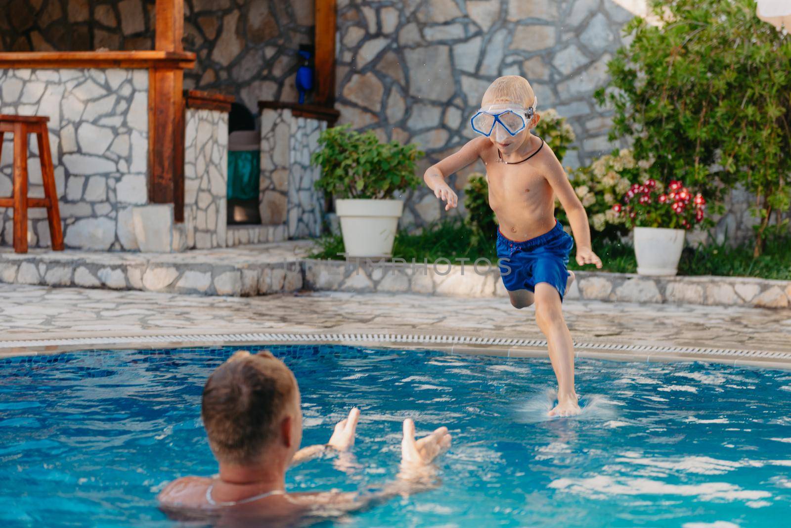 Excited boy in googles jumping in water from shoulders of his father standing in swimming pool by Andrii_Ko