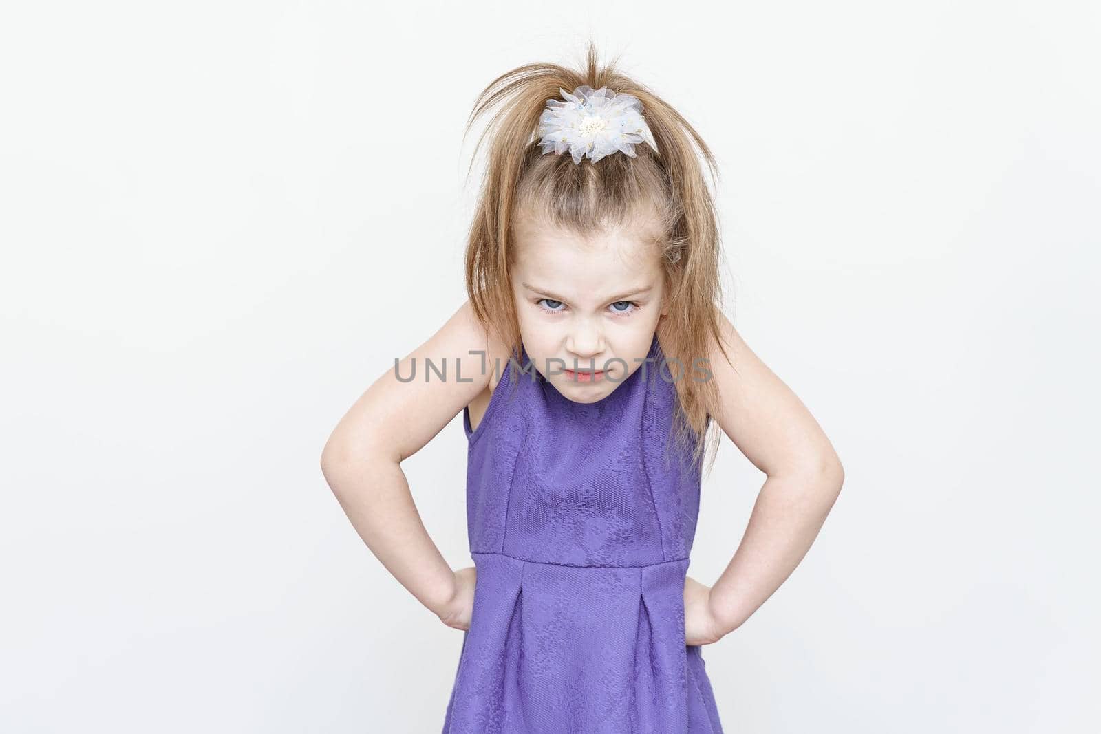 cute upset little girl in a blue dress. hands at sides, evil emotion. gray background