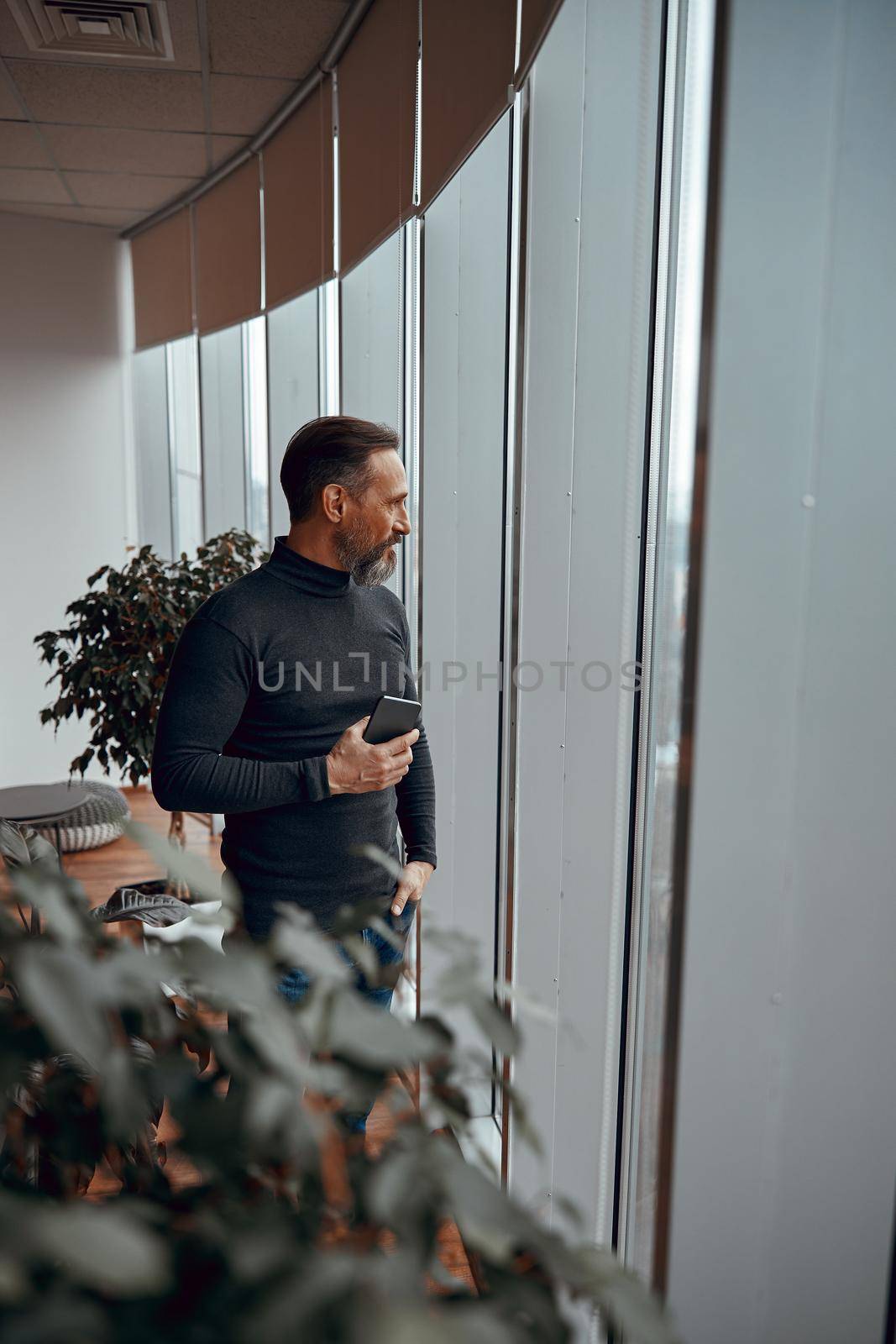 Calm man holding hia mobile phone and looking outdoors in his office