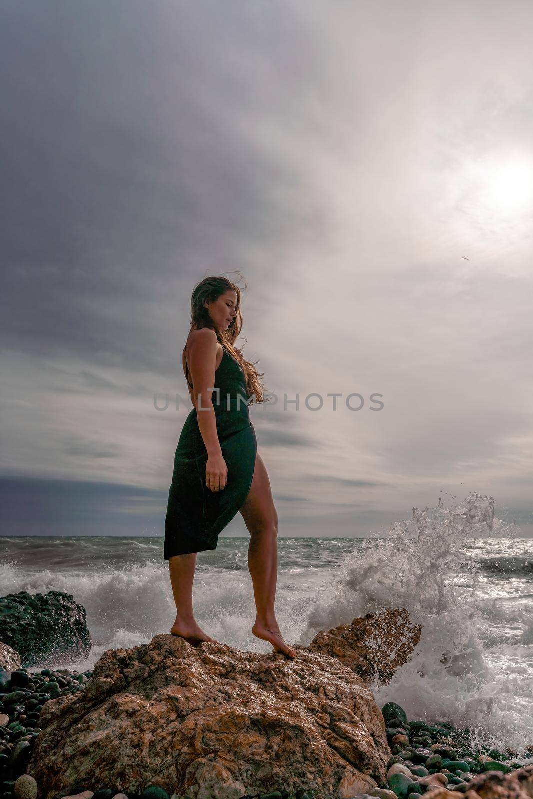 A beautiful girl in a black dress stands on a rock, big waves with white foam. A cloudy stormy day at sea, with clouds and big waves hitting the rocks. by Matiunina