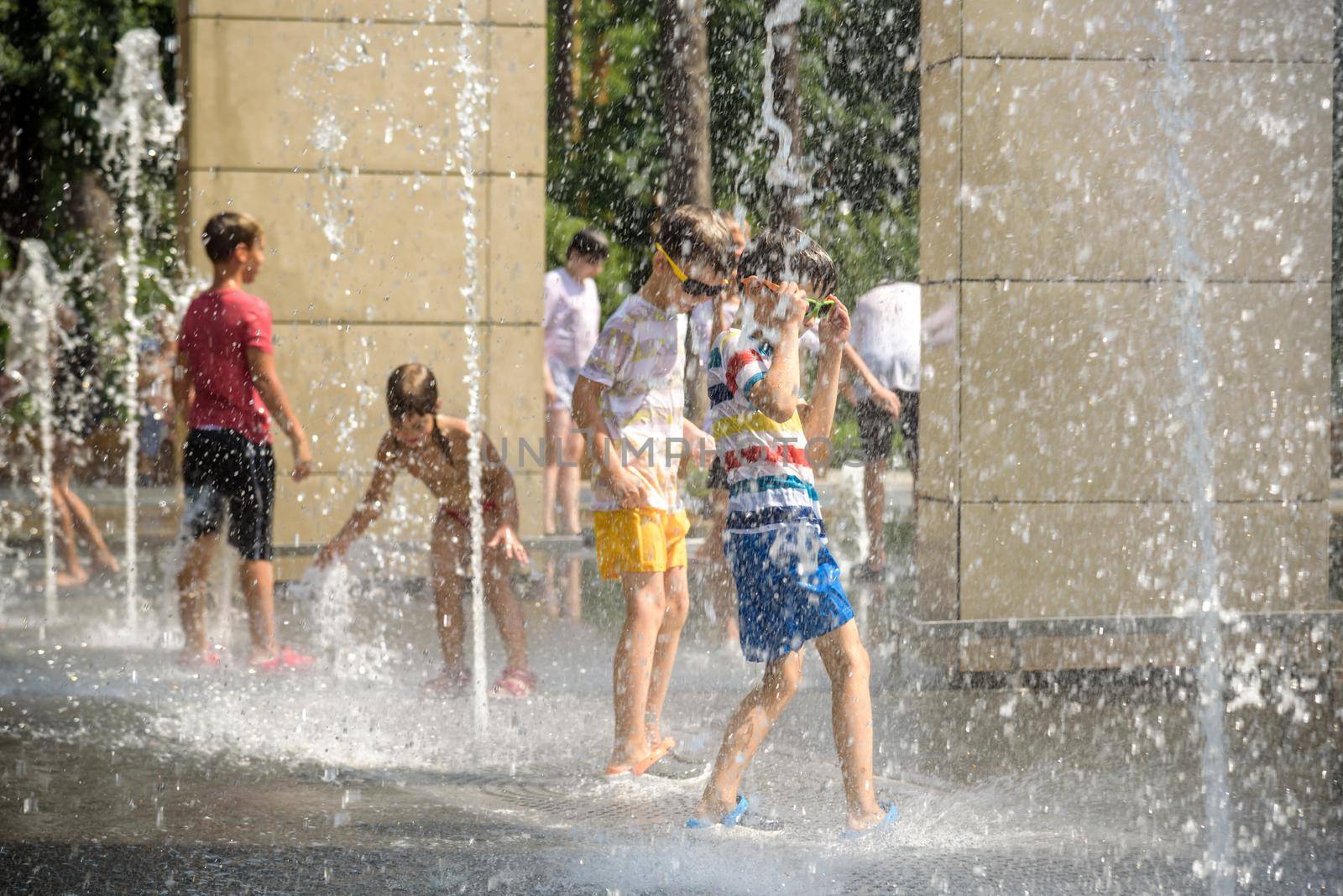 Kyiv, Ukraine - August 01, 2021: Boys jumping in water fountains. Children playing with a city fountain on hot summer day. Happy friends having fun in fountain. Summer weather. Friendship, lifestyle and vacation.