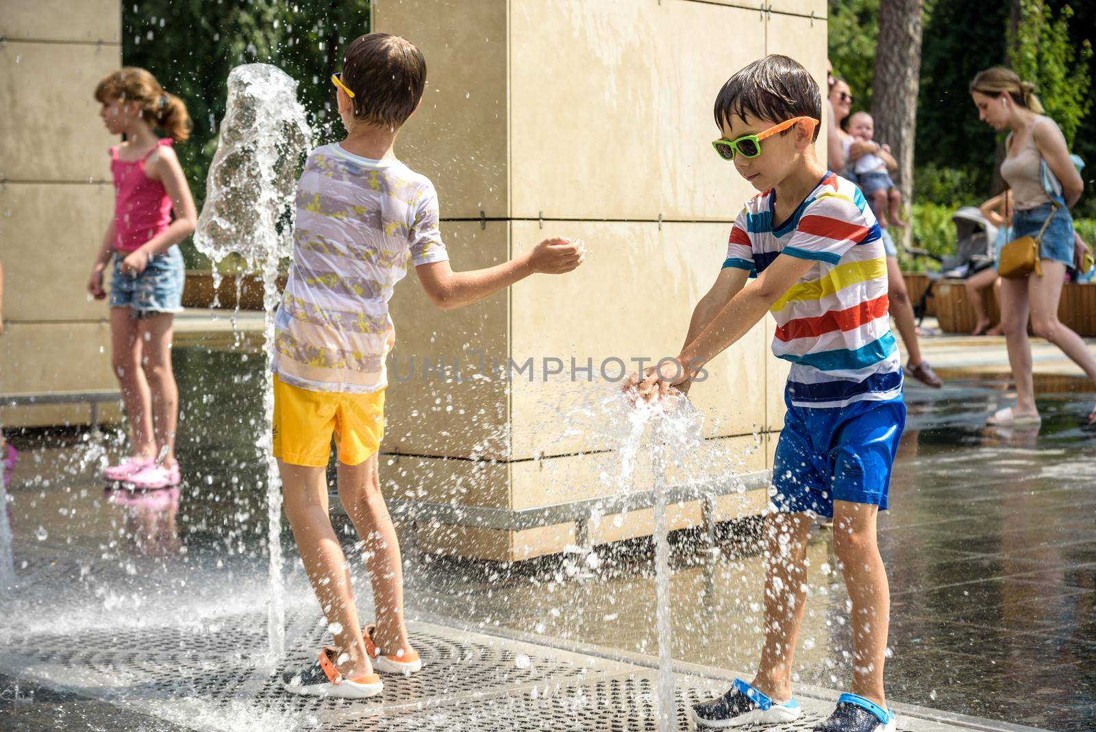 Kyiv, Ukraine - August 01, 2021: Boys jumping in water fountains. Children playing with a city fountain on hot summer day. Happy friends having fun in fountain. Summer weather. Friendship, lifestyle and vacation.