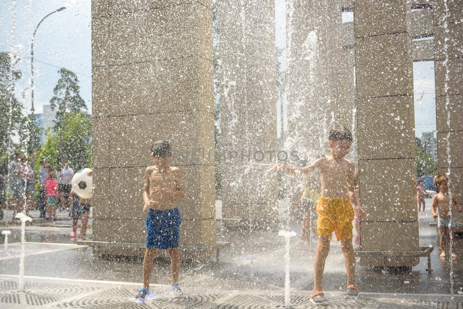 Kyiv, Ukraine - August 01, 2021: Boys jumping in water fountains. Children playing with a city fountain on hot summer day. Happy friends having fun in fountain. Summer weather. Friendship, lifestyle and vacation.