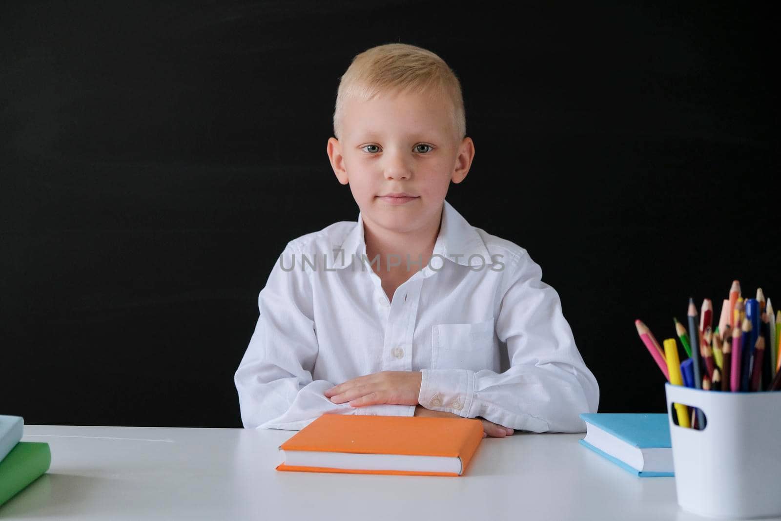 Cute little boy sitting at the table on blackboard backgrpund. Child from elementary school. Education concept.
