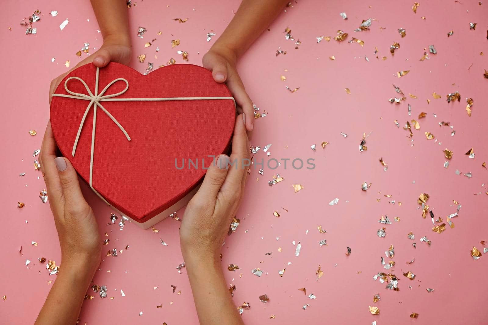 Red heart shape gift box on festive pink background, with child's and mom's hands. Mothers day mock up