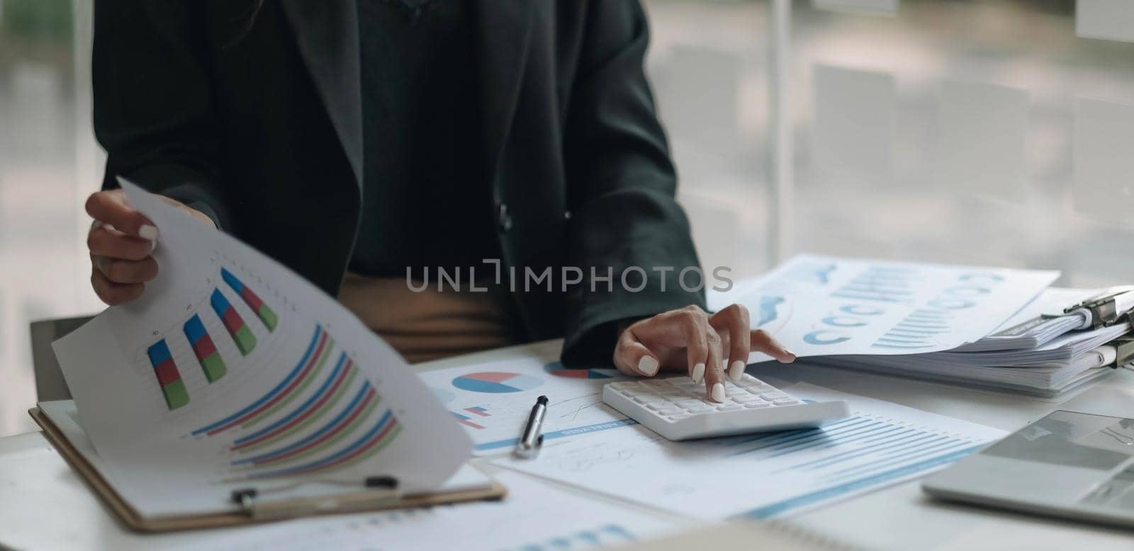 Close up of businesswoman or accountant hand holding pen working on calculator to calculate business data, accountancy document and laptop computer at office, business concept by wichayada