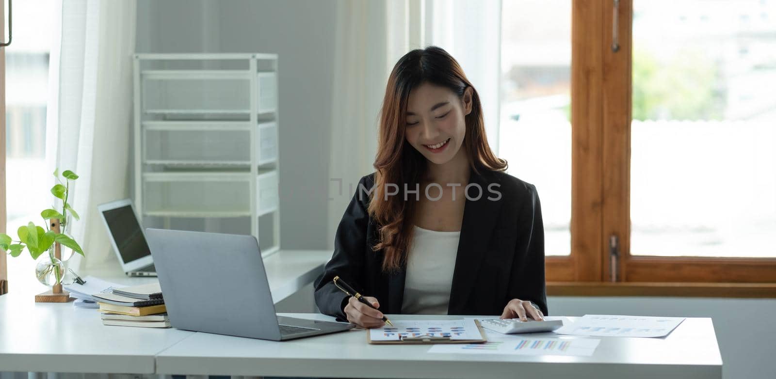 Beautiful Asian woman sitting in office using laptop and calculator. Happy business woman sitting at a desk in an office with a tablet computer.
