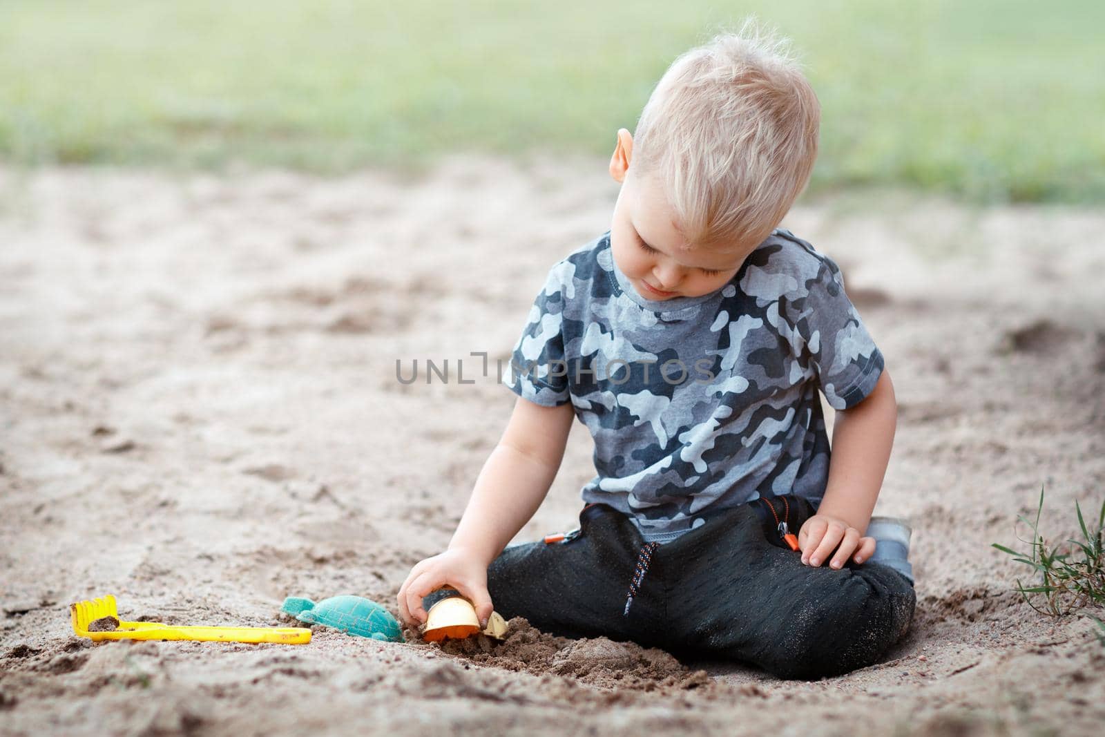 Alone child is sitting on a sand and calm playing with sand toys. Boy wearing military camouflage shirts playing on a ground in outdoor