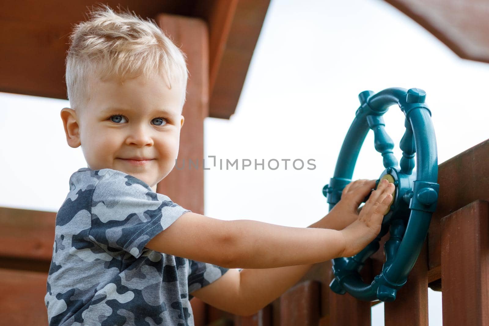 Happy child is driving a ship wheel in a playground. Kid wearing camouflage is making a signal for boats in a playhouse. Beeping horn. Childhood full of adventure