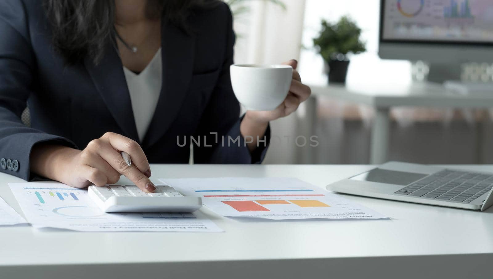 Asian businesswoman working on laptop computer and drinking coffee in modern office near window with calculator and business report on desk. work from home concept