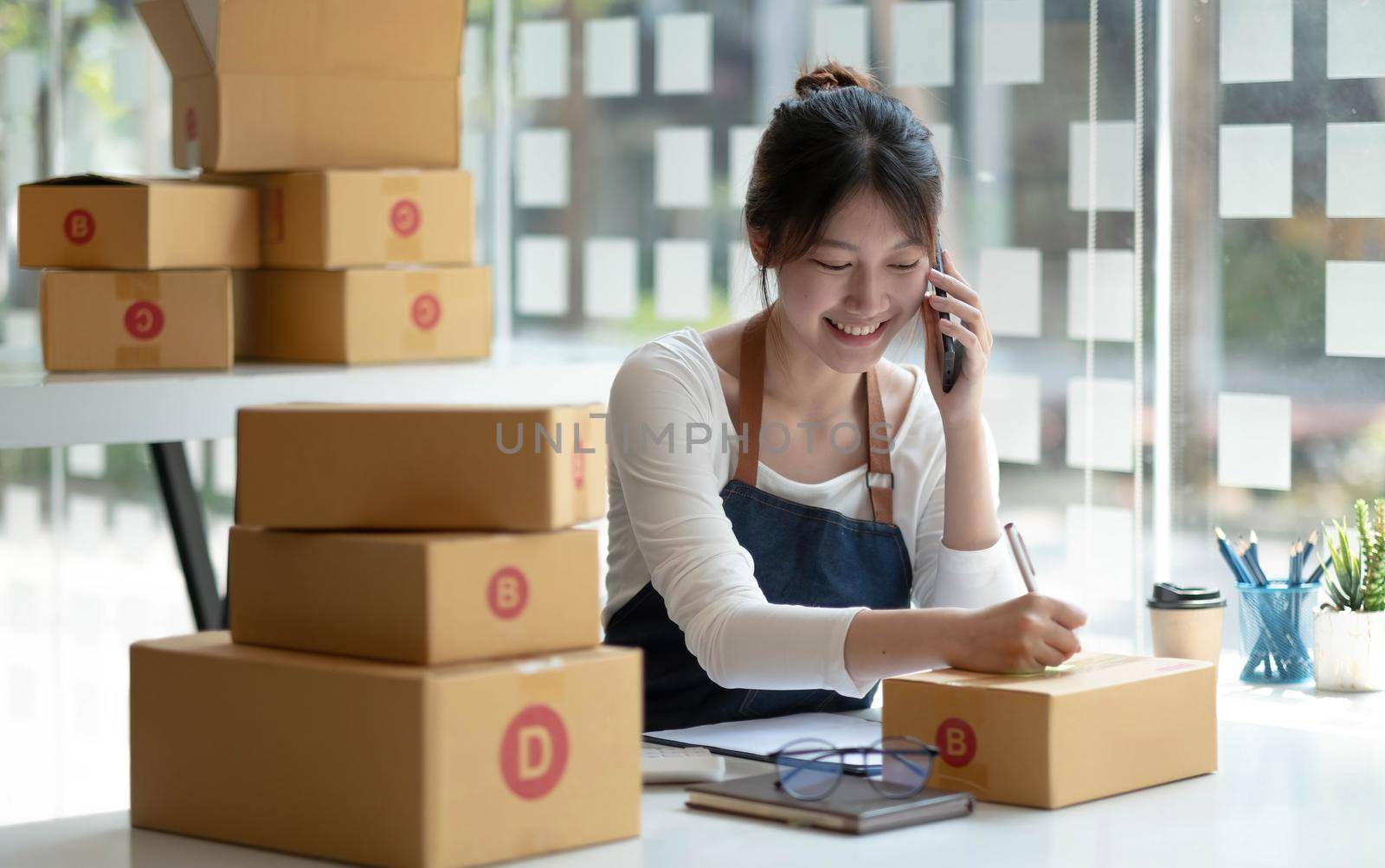 Happy young Asian business owner woman prepare parcel box and check online orders of product for deliver to customer on smartphone. Shopping Online concept. by wichayada
