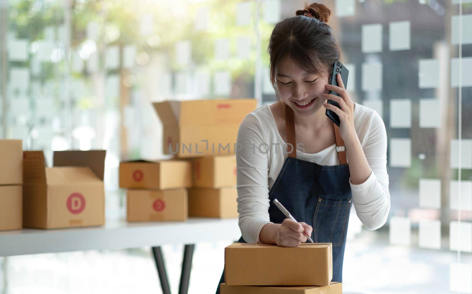 Happy young Asian business owner woman prepare parcel box and check online orders of product for deliver to customer on smartphone. Shopping Online concept..